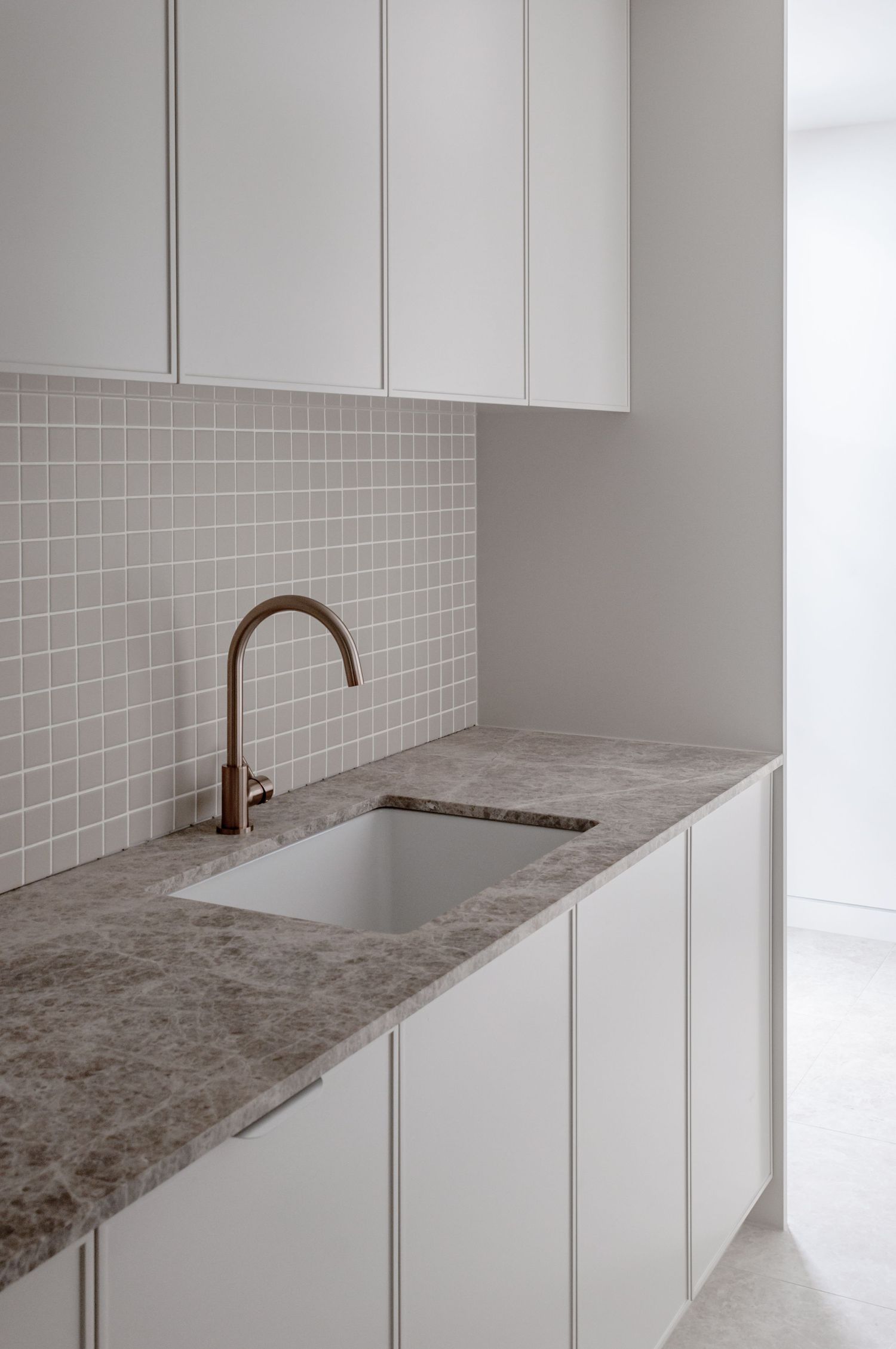 Minimalist laundry room with white cabinets, gray countertop and brass faucet against tiled backsplash.