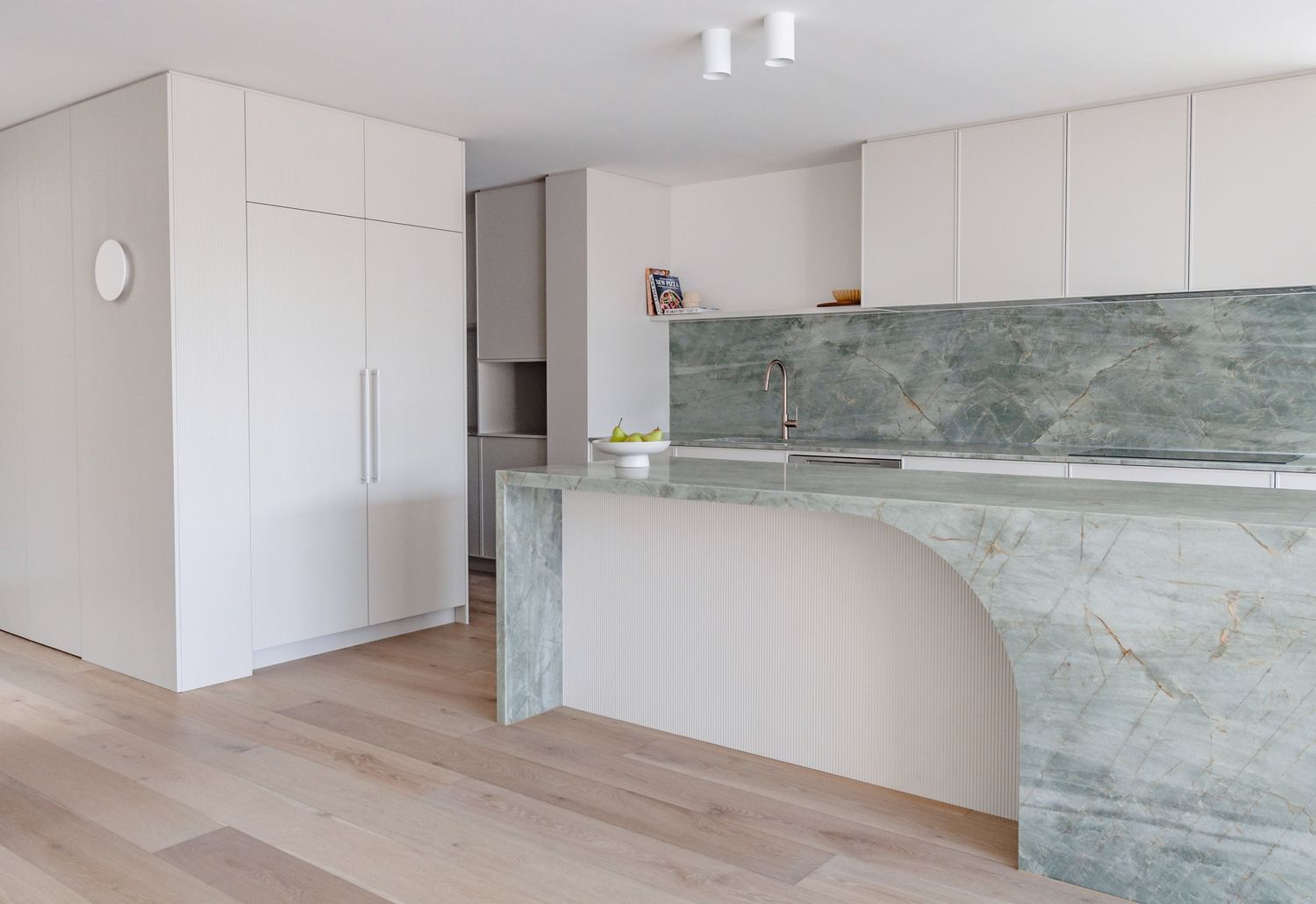 Modern minimalist kitchen featuring white cabinets and a curved green marble island with sleek lighting fixtures.