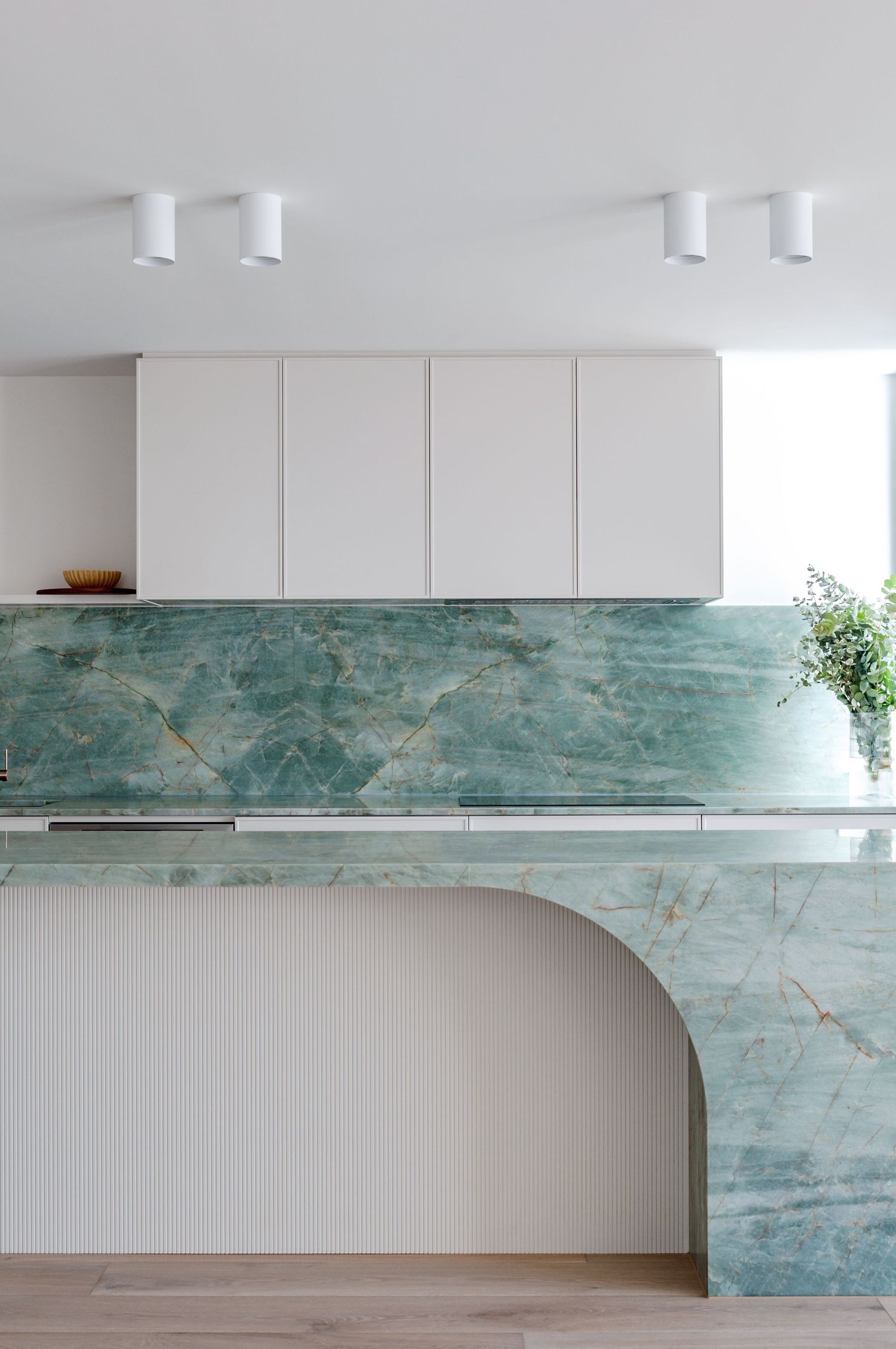 Close-up of elegant green marble backsplash with white upper cabinets and cylindrical ceiling lights.