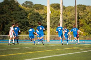 Soccer players sit on a bench during a sunny day match with trees in the background.
