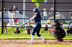 Players on a softball field with chain link fence and trees in background.