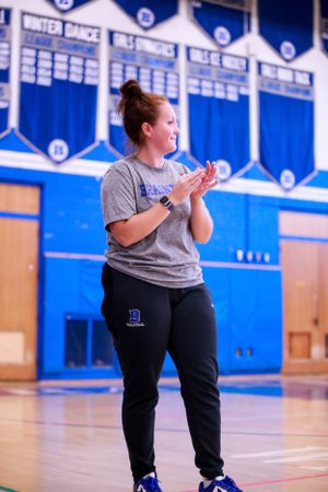 Coach demonstrates volleyball techniques in a blue and white school gymnasium during practice.