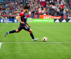 Soccer action shot shows players diving and sliding on green turf during competitive match.