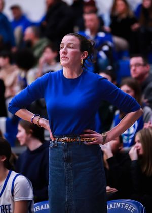 Coach in blue sweater and jeans stands on sideline during basketball game.