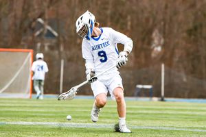 Lacrosse players in white and navy uniforms battle for control during a competitive game.