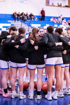 Basketball team stands in unity with arms around each other wearing matching uniforms.