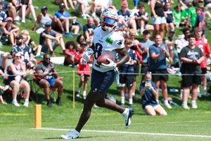 Players run football drills as spectators watch from the sidelines on a grassy field.