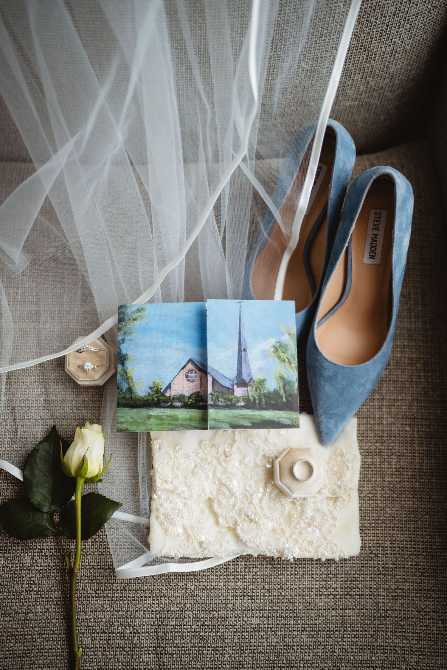 Wedding details arranged on gray linen including blue shoes, white veil, rose flower, and scenic photo cards.