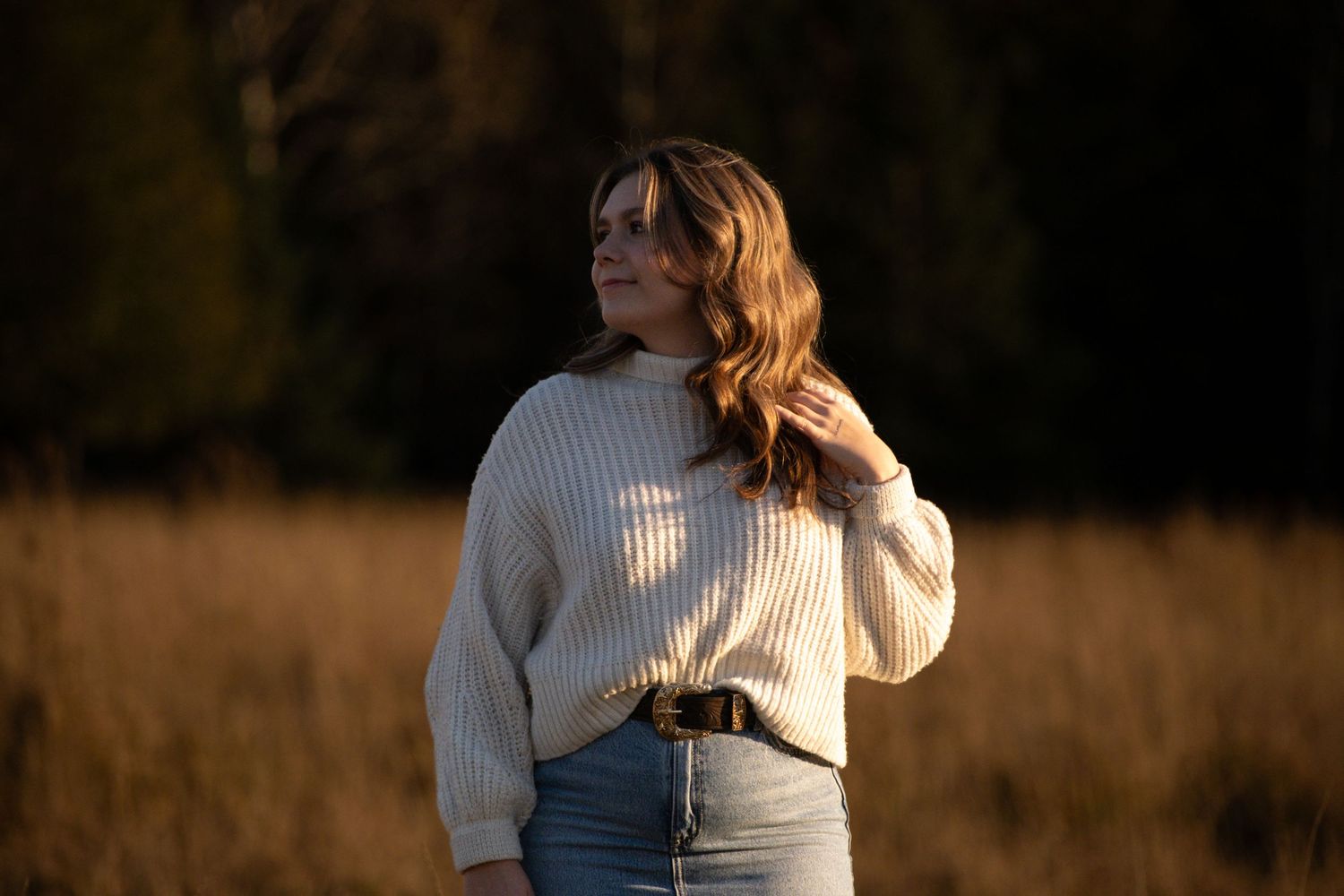 Person in cream sweater standing in golden sunlit field at sunset.