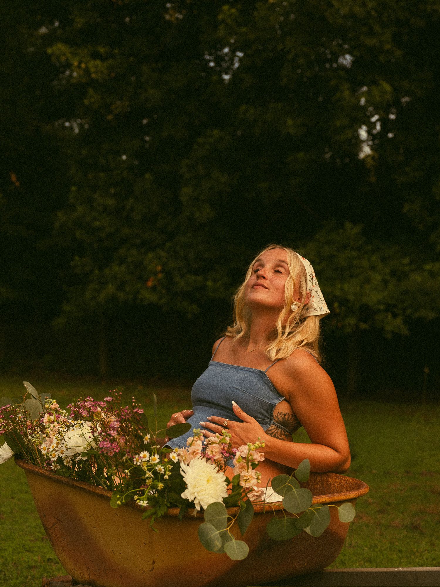 Pregnant woman in blue top sits in rustic wheelbarrow filled with flowers, gazing upward in lush green garden.