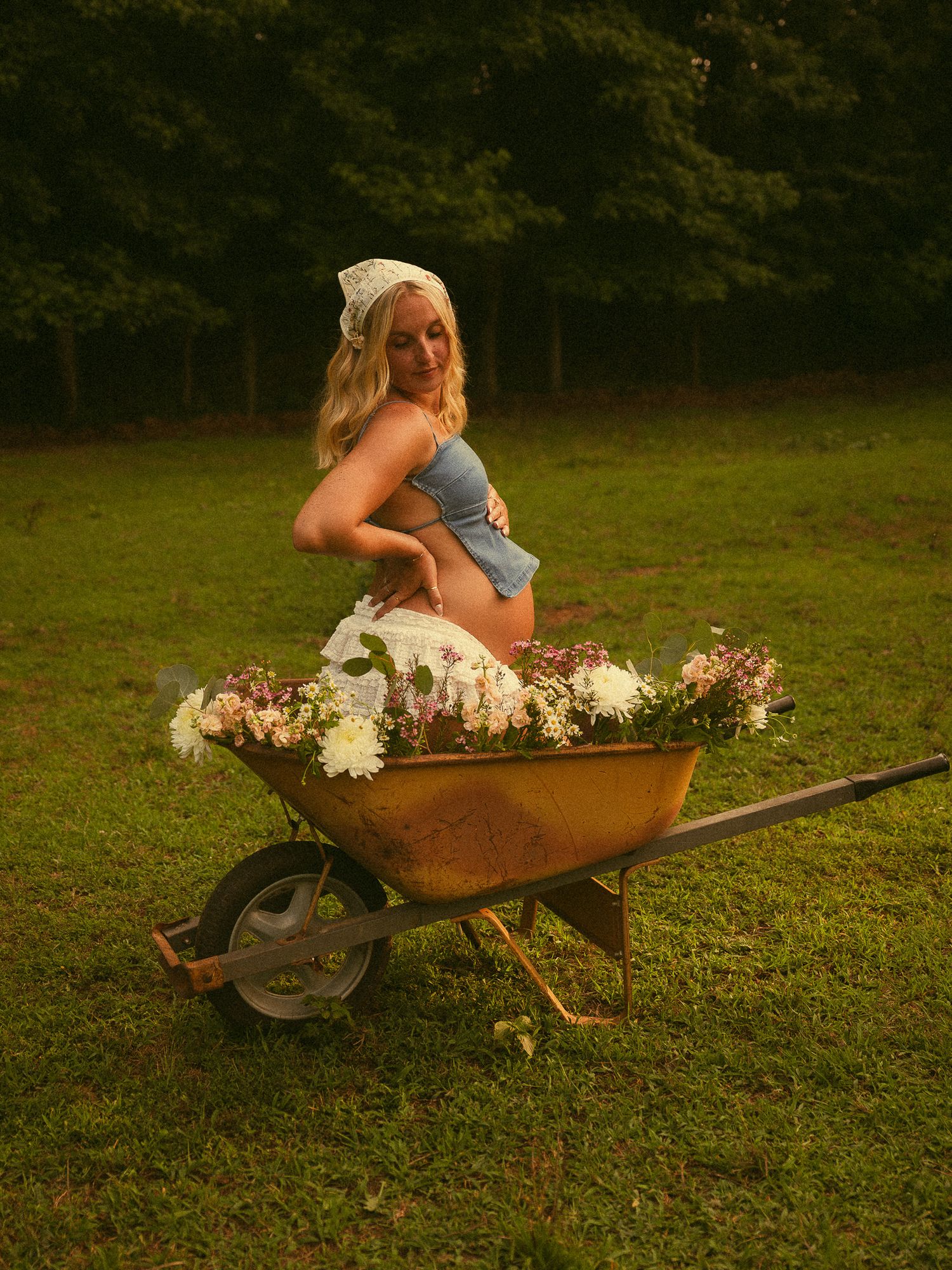 Pregnant woman sitting in a rustic wheelbarrow filled with flowers on green grass, wearing a blue top and white headscarf.