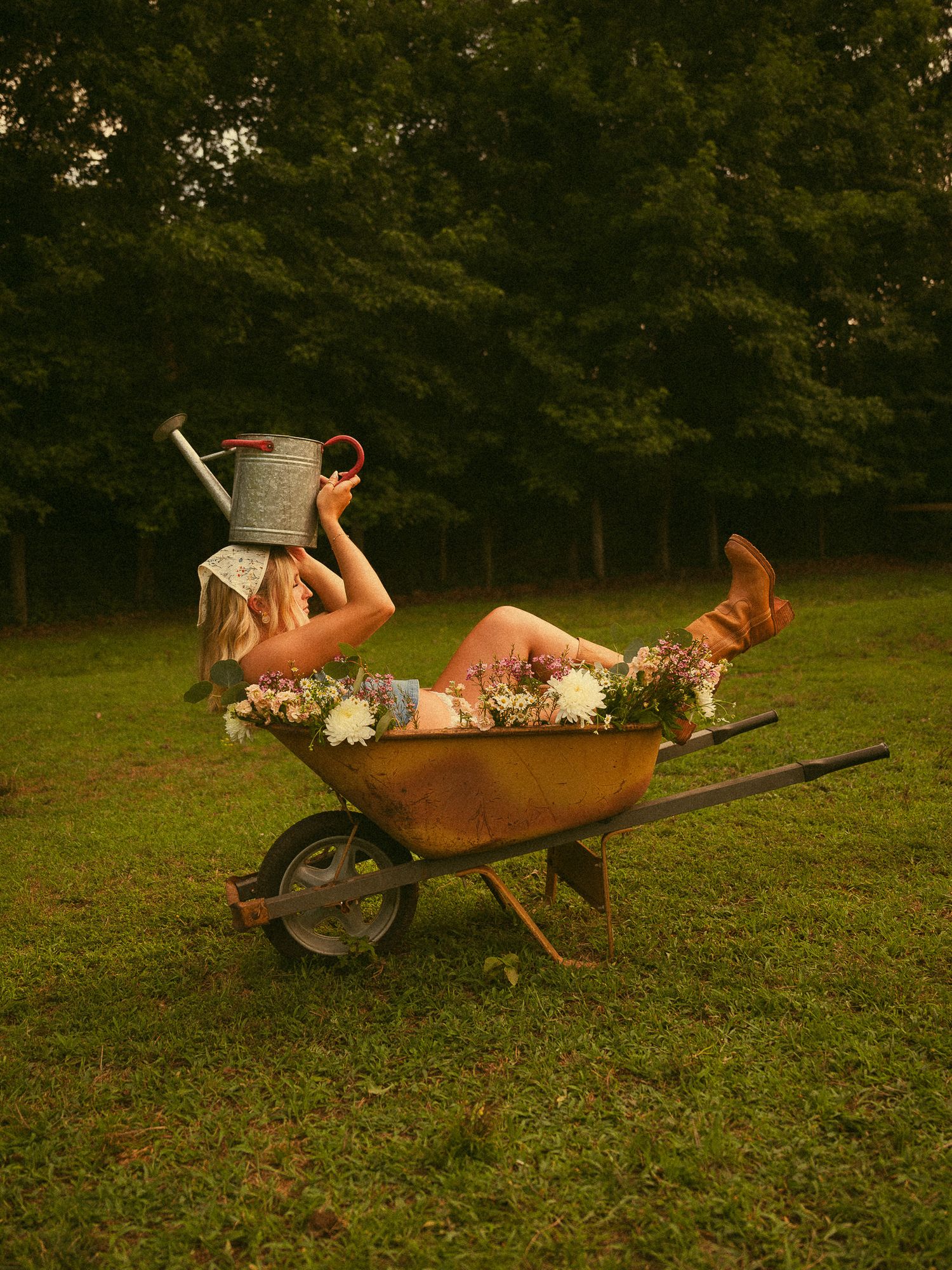 A woman reclines in a rustic wheelbarrow filled with flowers, holding a watering can, surrounded by lush green trees.