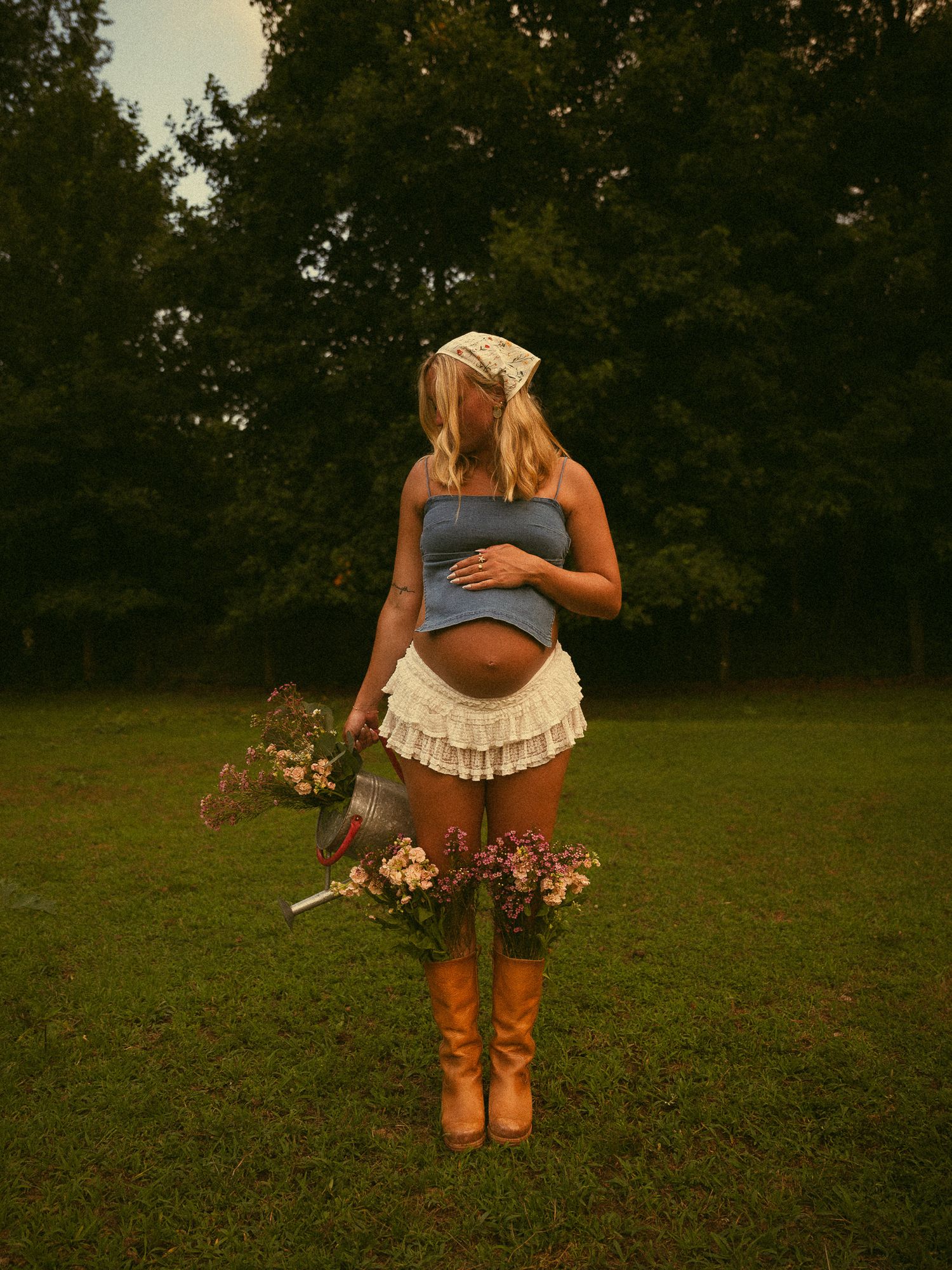 Pregnant woman in blue crop top, ruffled skirt, and brown boots holding wildflowers outdoors in a grassy field.