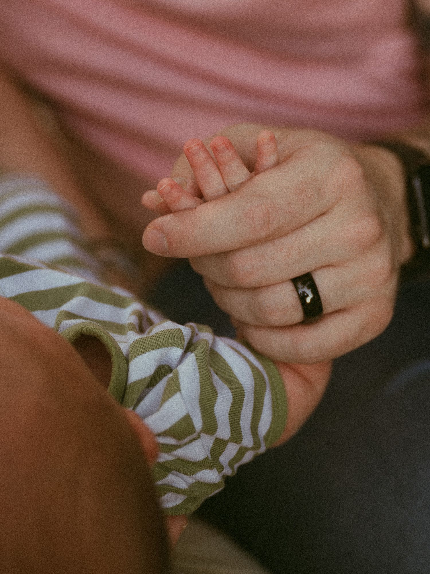 A parent with a wedding ring gently holds a newborn baby's tiny hand, dressed in a green striped onesie.