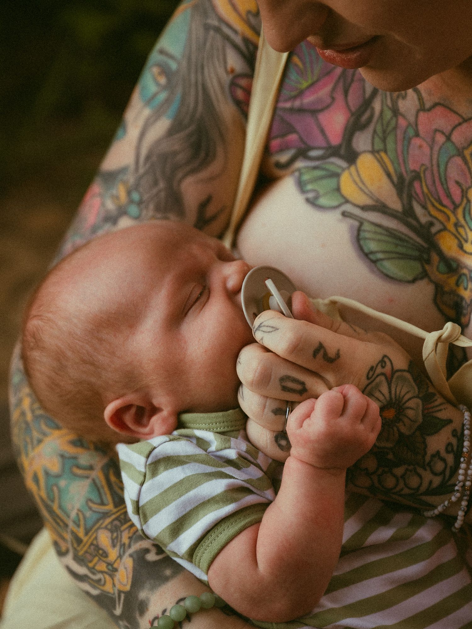 Heavily tattooed mother breastfeeding her newborn baby outdoors, colorful floral tattoos covering her arms and chest.
