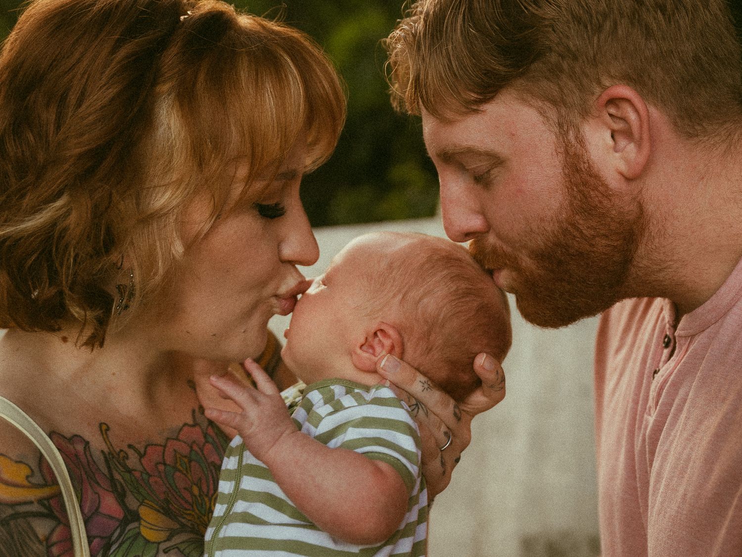 New parents kissing their newborn baby together, mother with red hair and tattoos, father with beard, outdoors.