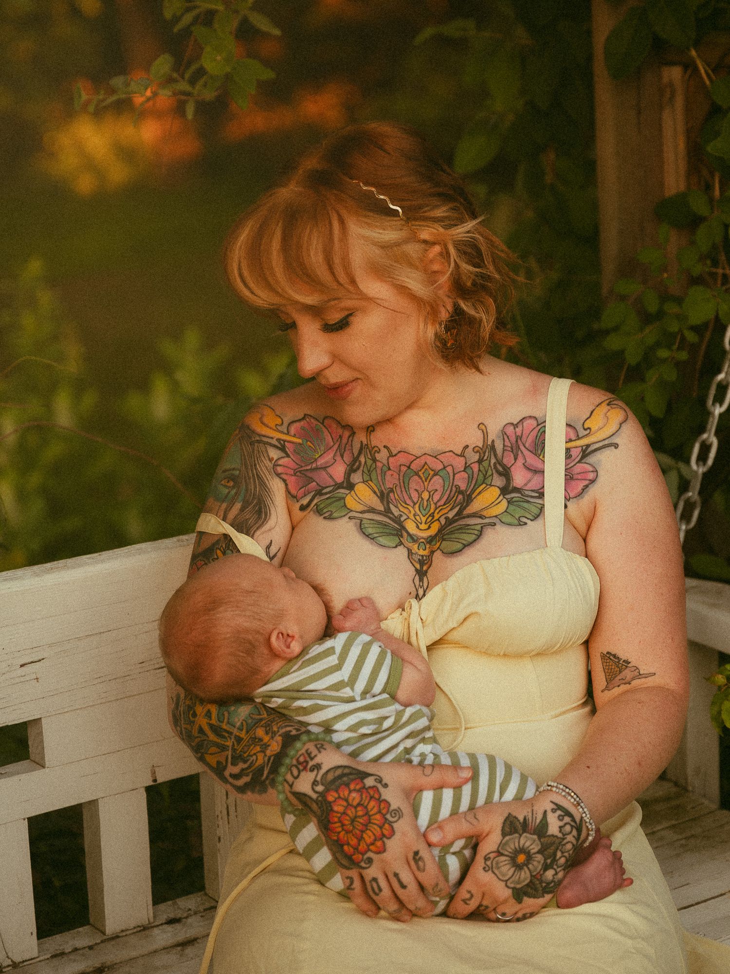 Tattooed mother breastfeeding newborn baby on white wooden bench in lush green garden setting.