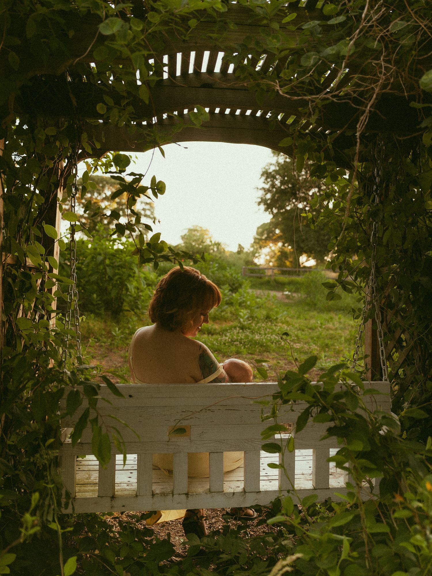 Child sitting on a white wooden bench swing framed by a lush green garden arbor at golden hour.