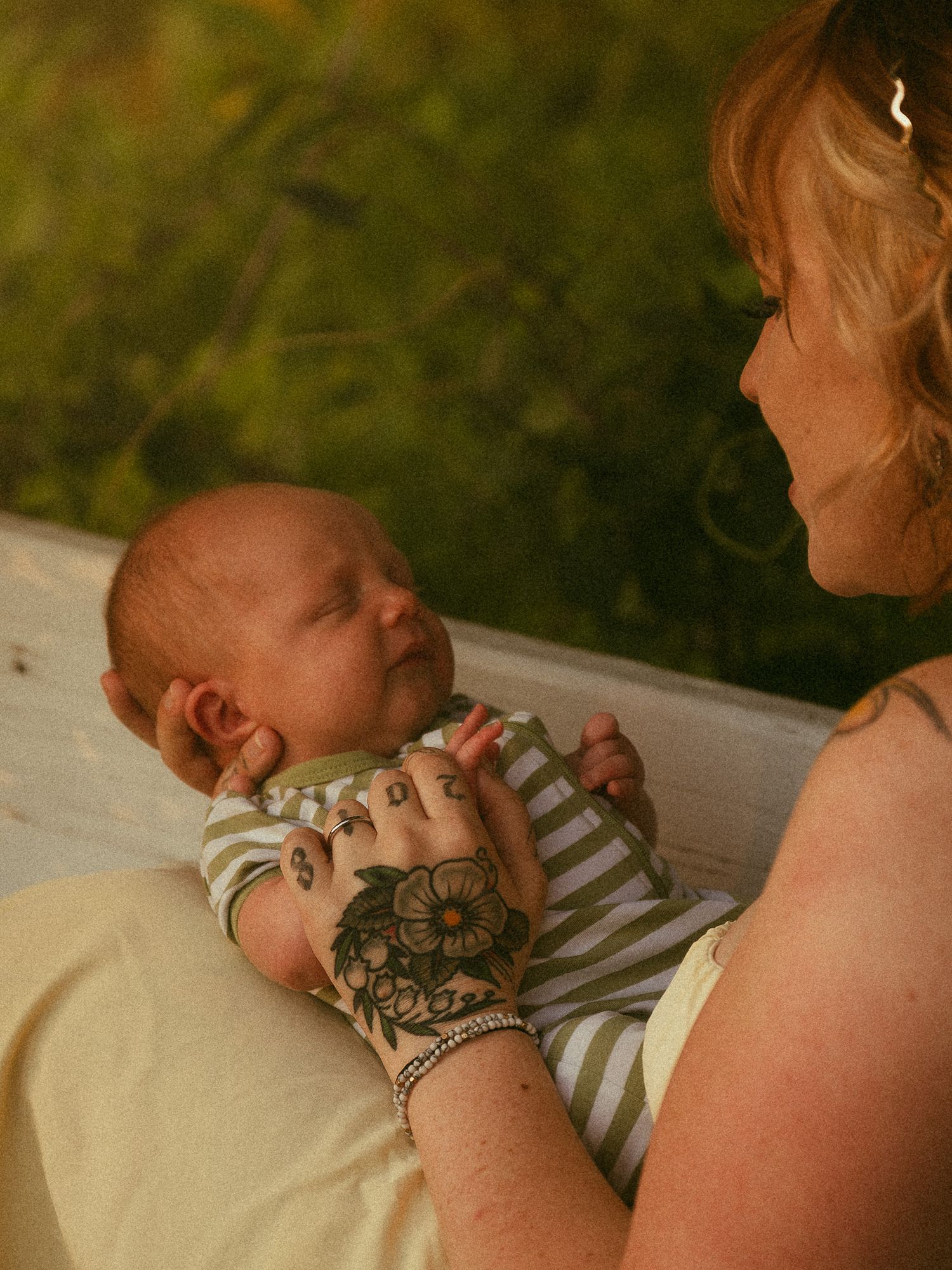 A mother with floral hand tattoos tenderly holds her newborn baby outdoors in soft golden light.