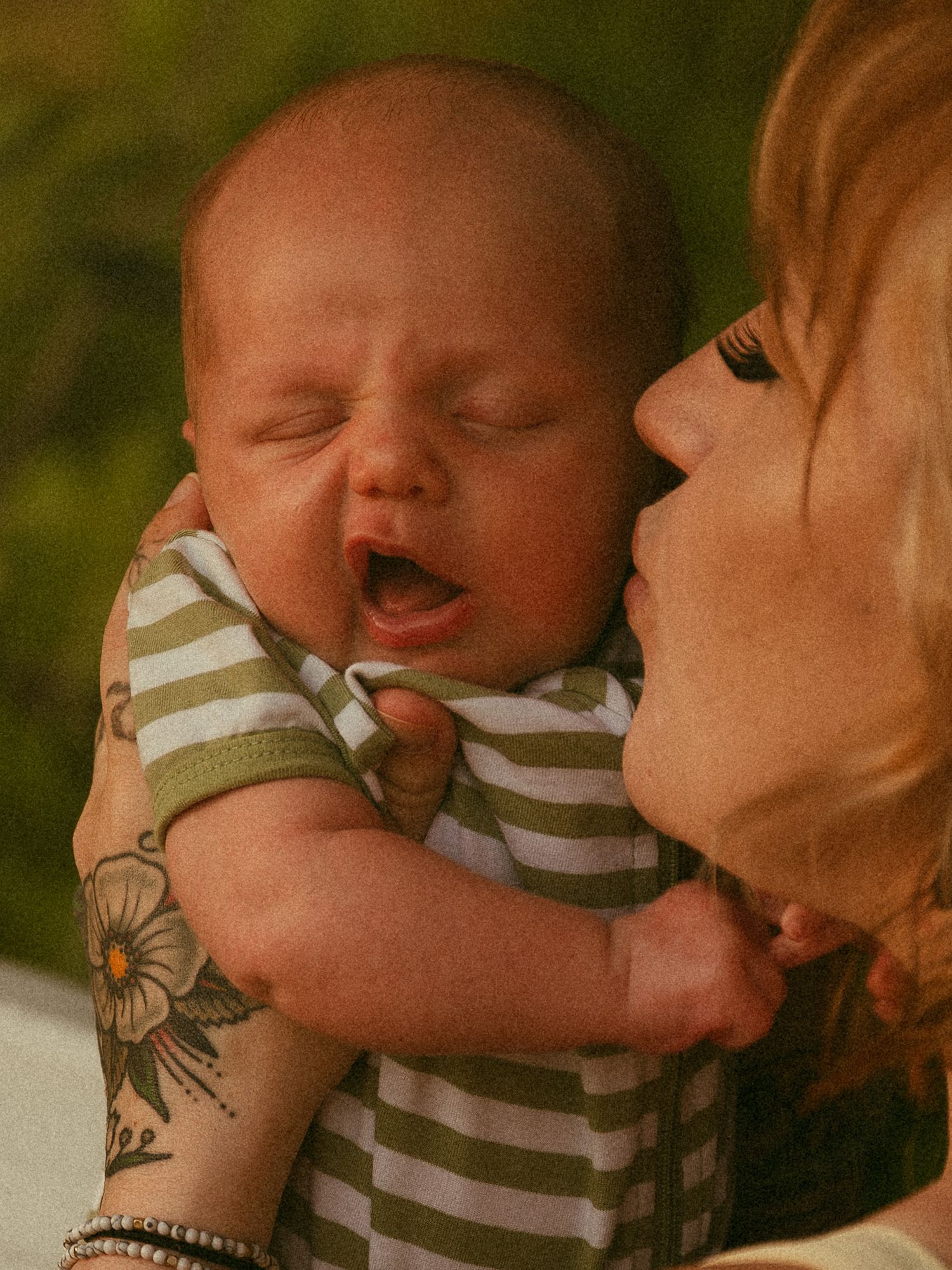 A tattooed mother holds her yawning newborn baby dressed in a green striped onesie outdoors.