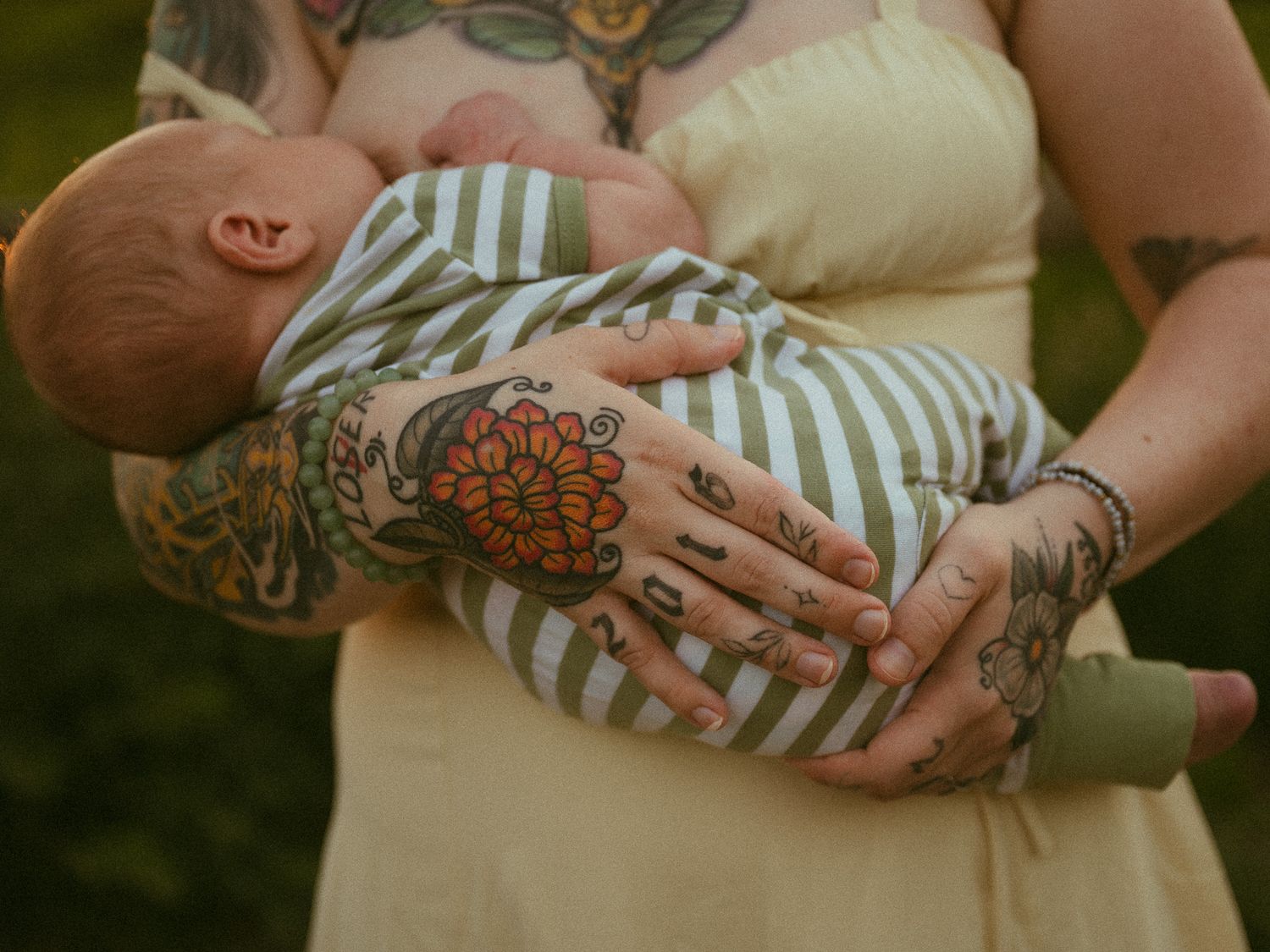 Tattooed mother cradling a newborn baby in her arms, wearing a yellow dress outdoors.
