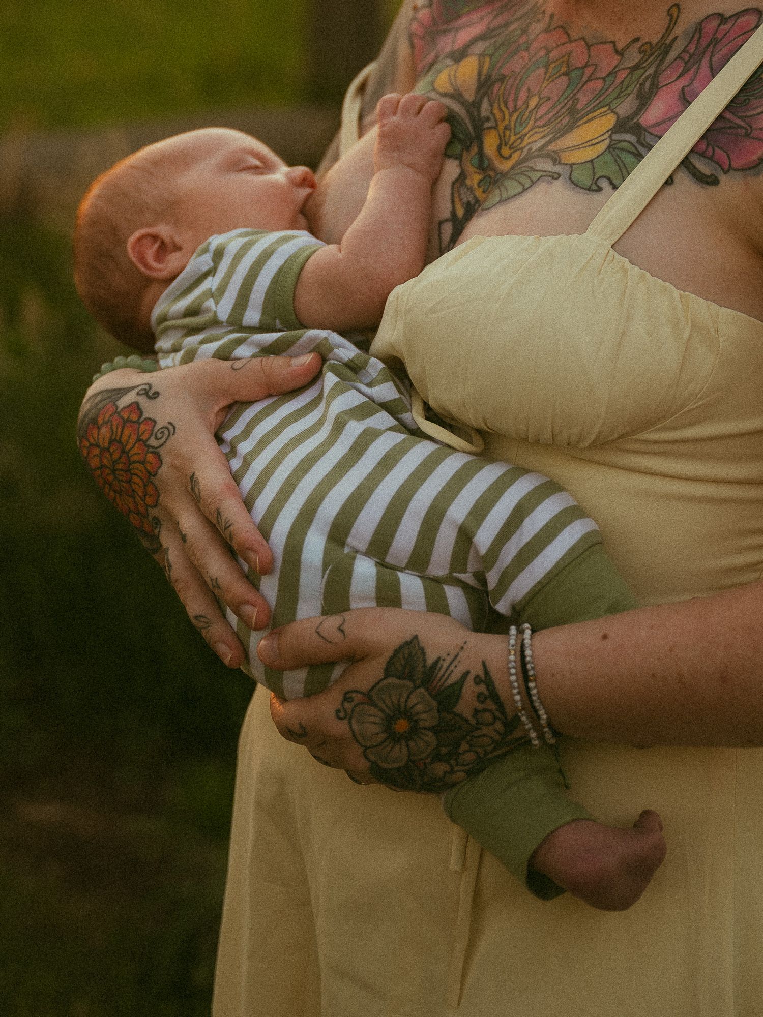 Tattooed mother holding newborn baby dressed in striped onesie outdoors in soft natural light.