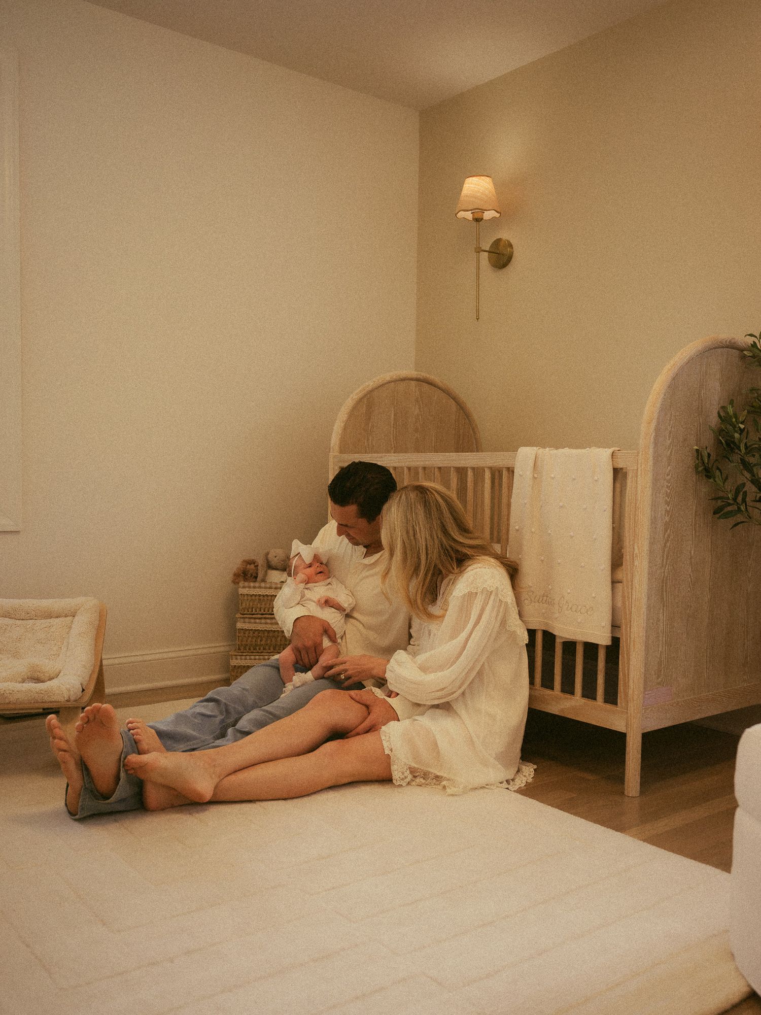 Couple sitting on nursery floor with newborn baby near wooden crib in soft neutral toned room.