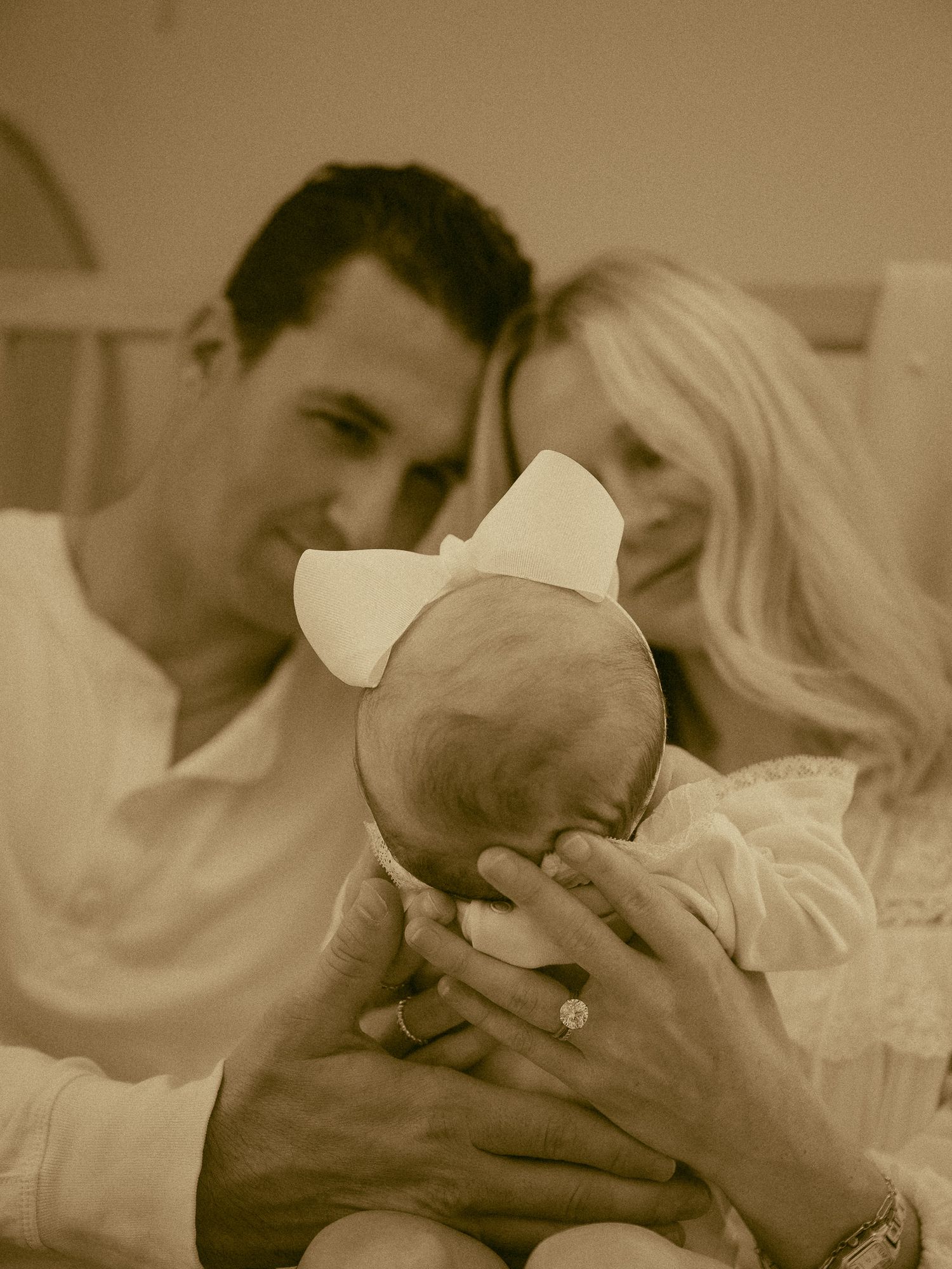 Sepia-toned portrait of parents tenderly holding newborn baby with white bow in intimate family moment.