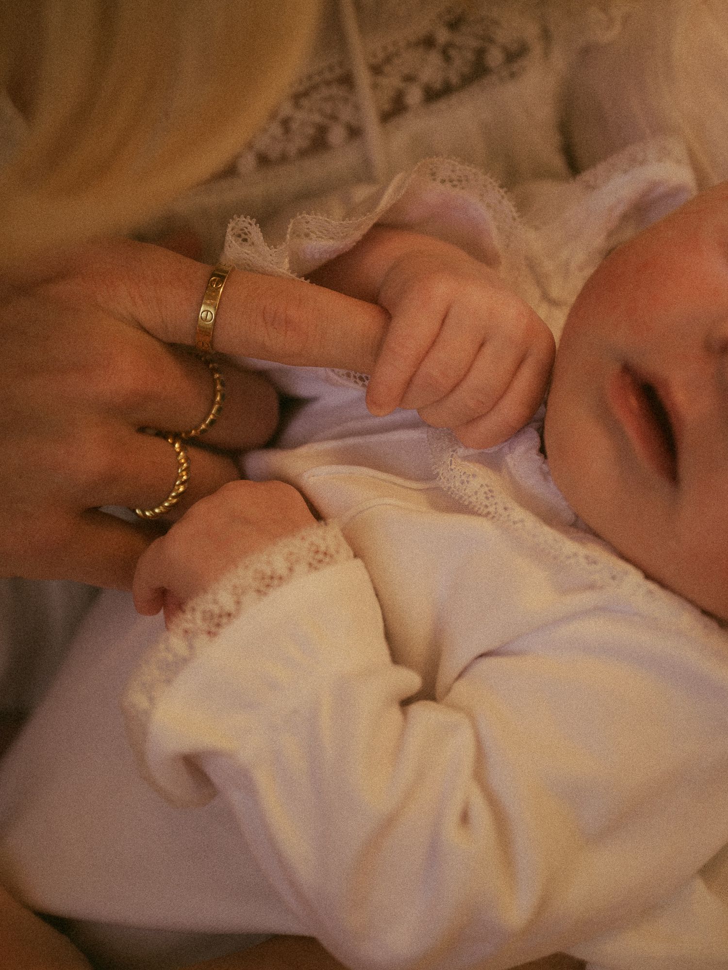 A sleeping newborn baby in white lace outfit holds an adult finger adorned with gold rings.