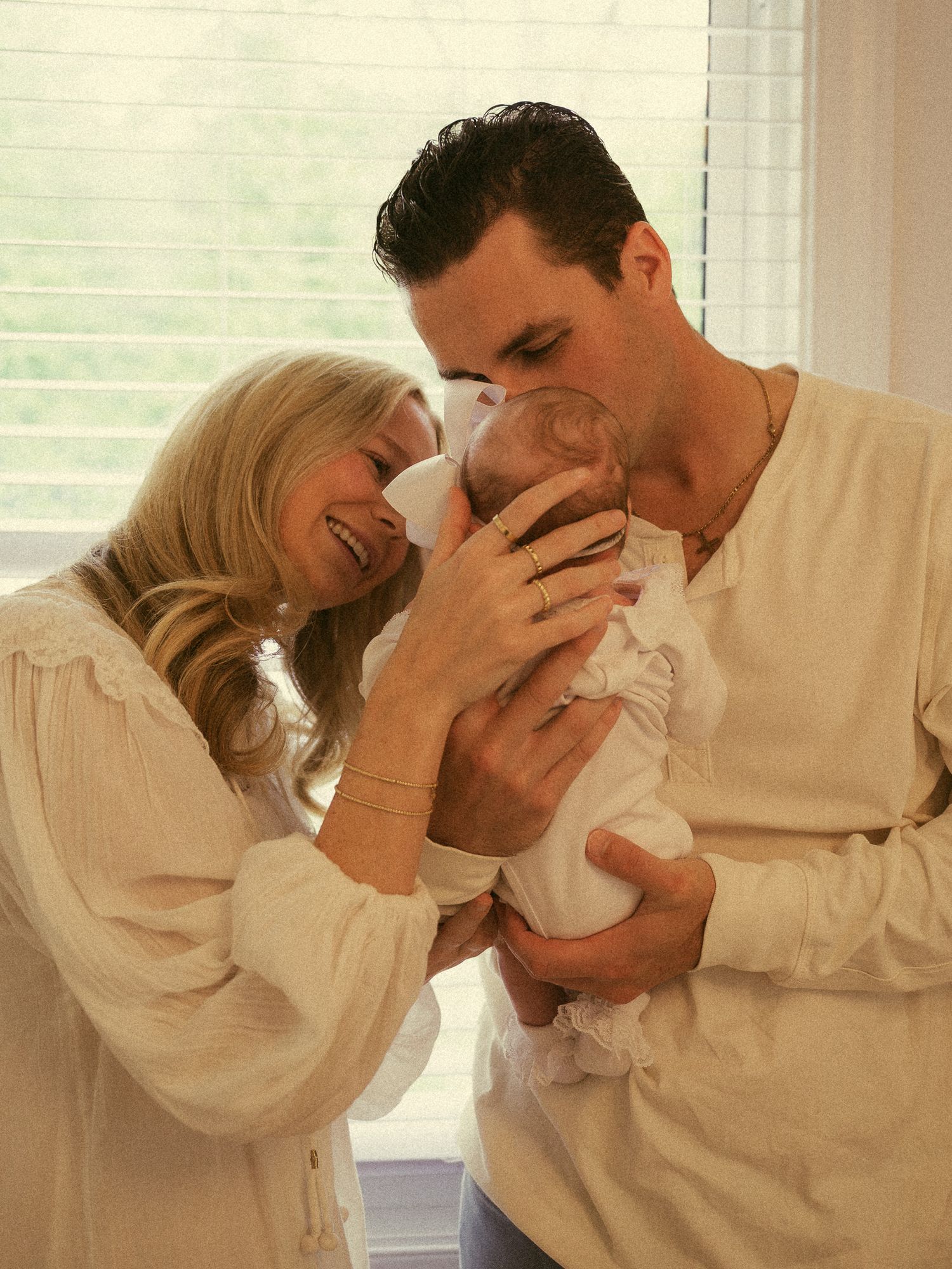 Parents lovingly kissing and cuddling newborn baby by bright window in warm intimate family portrait.