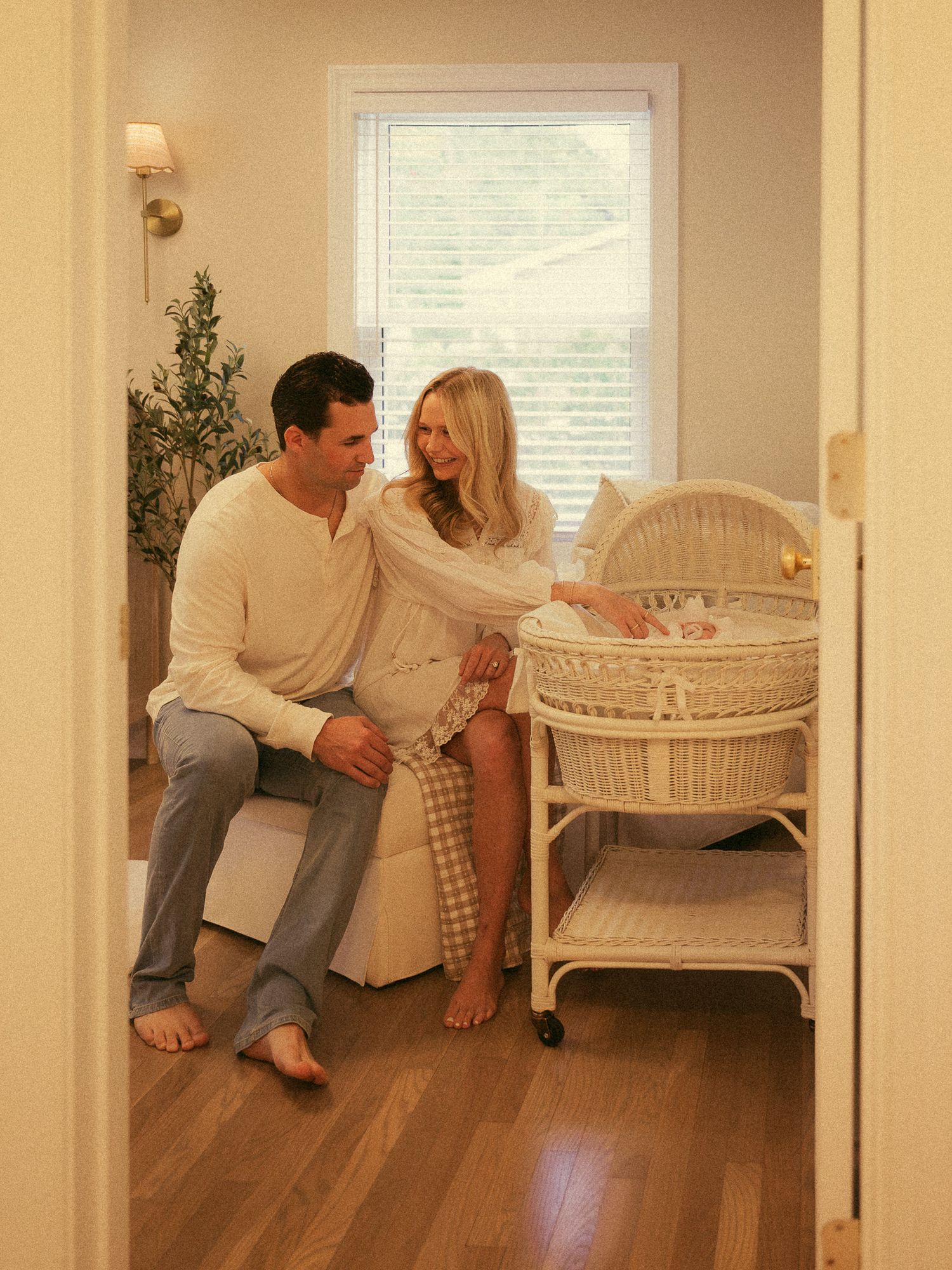 A couple sits together in a cozy nursery, smiling at their newborn baby in a white wicker bassinet.