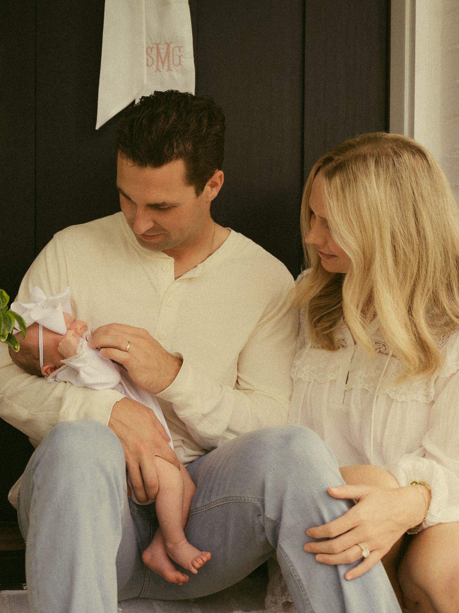 A couple sits on porch steps lovingly gazing at their newborn baby wrapped in white with a pink bow.
