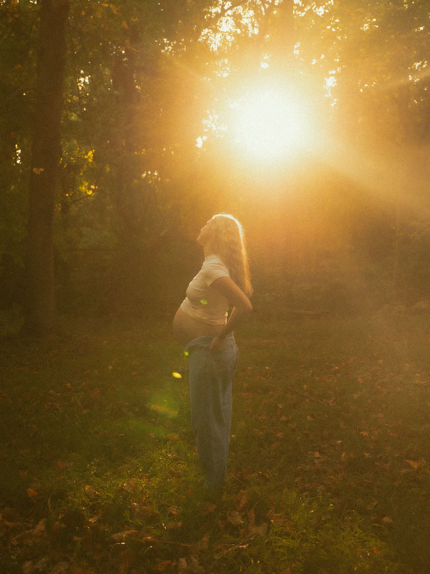 Pregnant woman standing in sunlit forest during golden hour, surrounded by autumn leaves and glowing warm light.