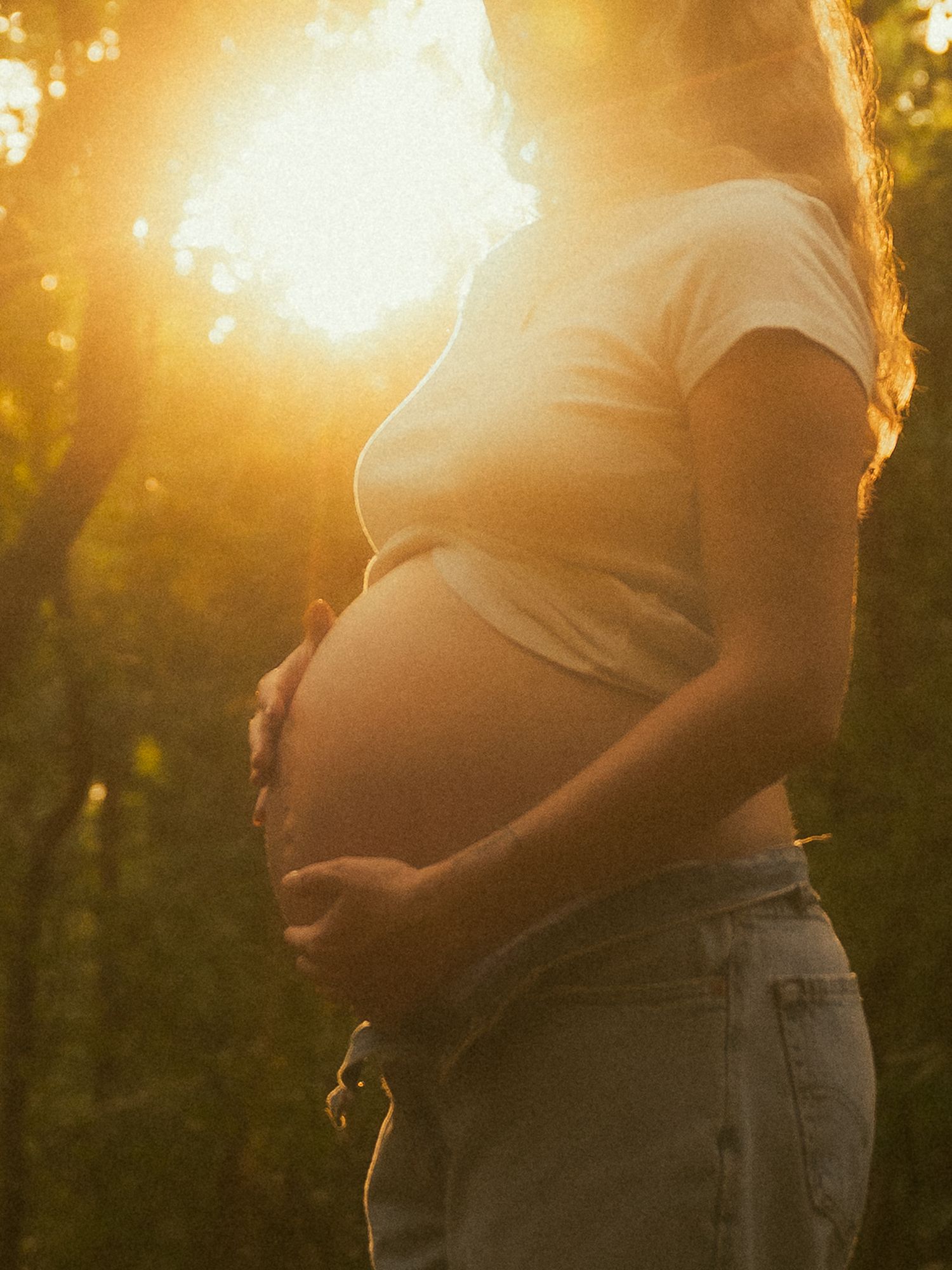 Pregnant woman cradling baby bump outdoors in golden sunset light wearing white t-shirt and jeans.