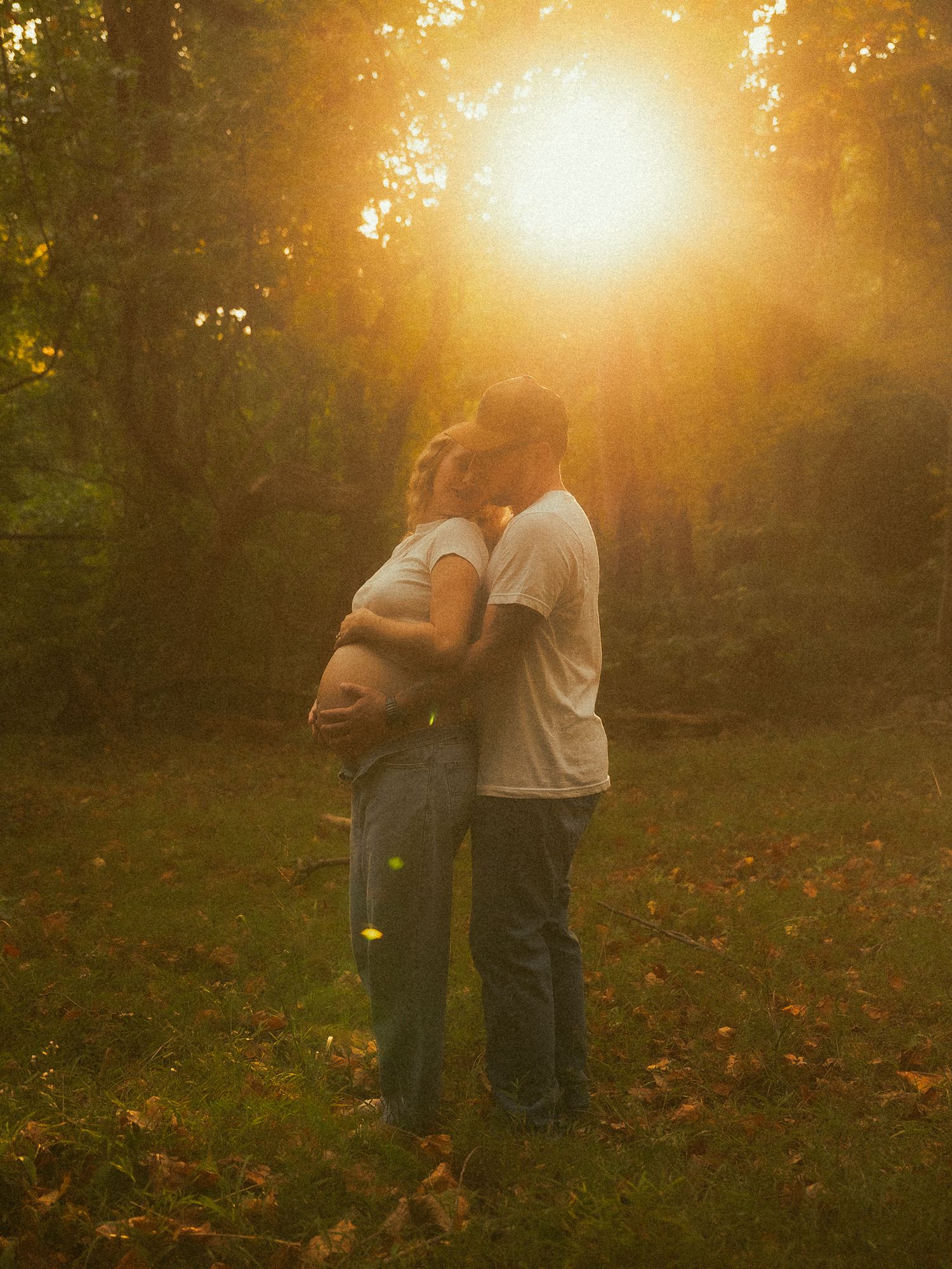 Couple sharing a tender kiss during golden hour sunset in a lush green forest, with woman cradling pregnant belly.