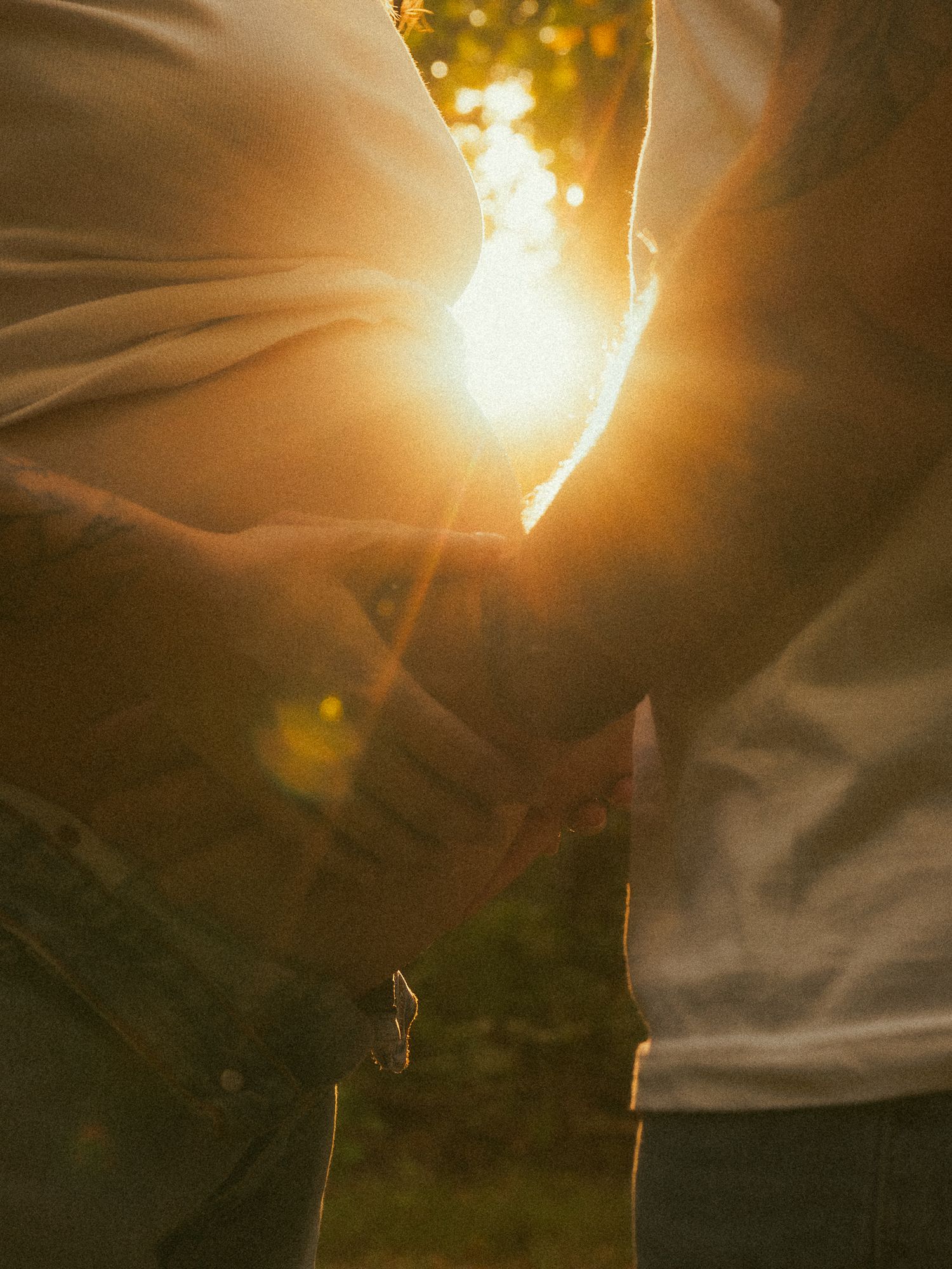 Two people holding hands at golden hour sunset with warm sunlight glowing between them outdoors.
