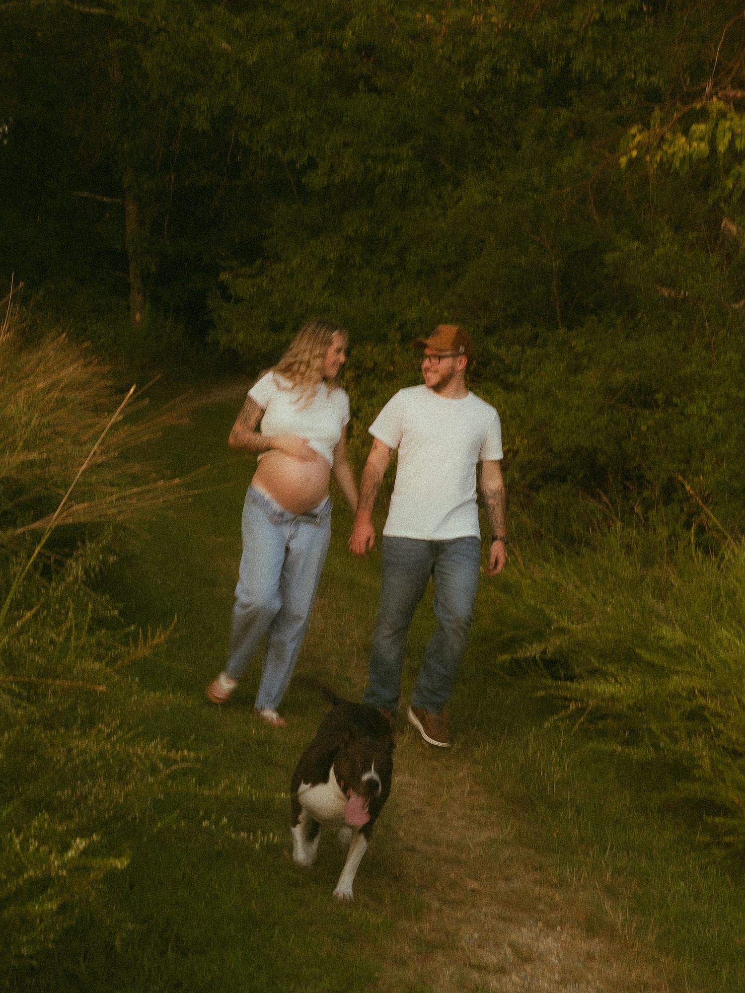 Pregnant couple holding hands walking on a nature trail with their dog amid lush green trees at golden hour.