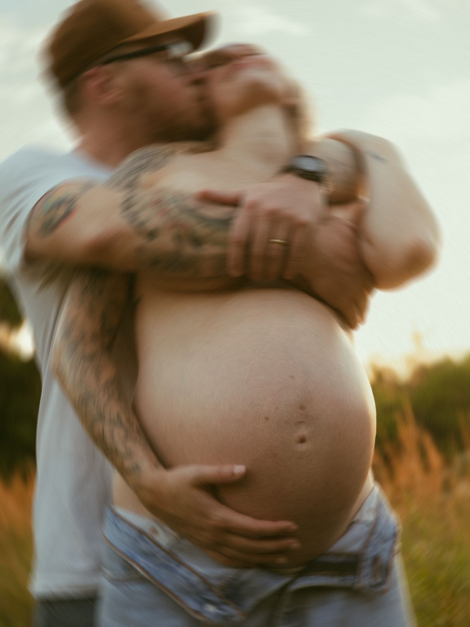 A tattooed man embraces his pregnant partner outdoors at sunset, gently cradling her bare baby bump in a tender moment.