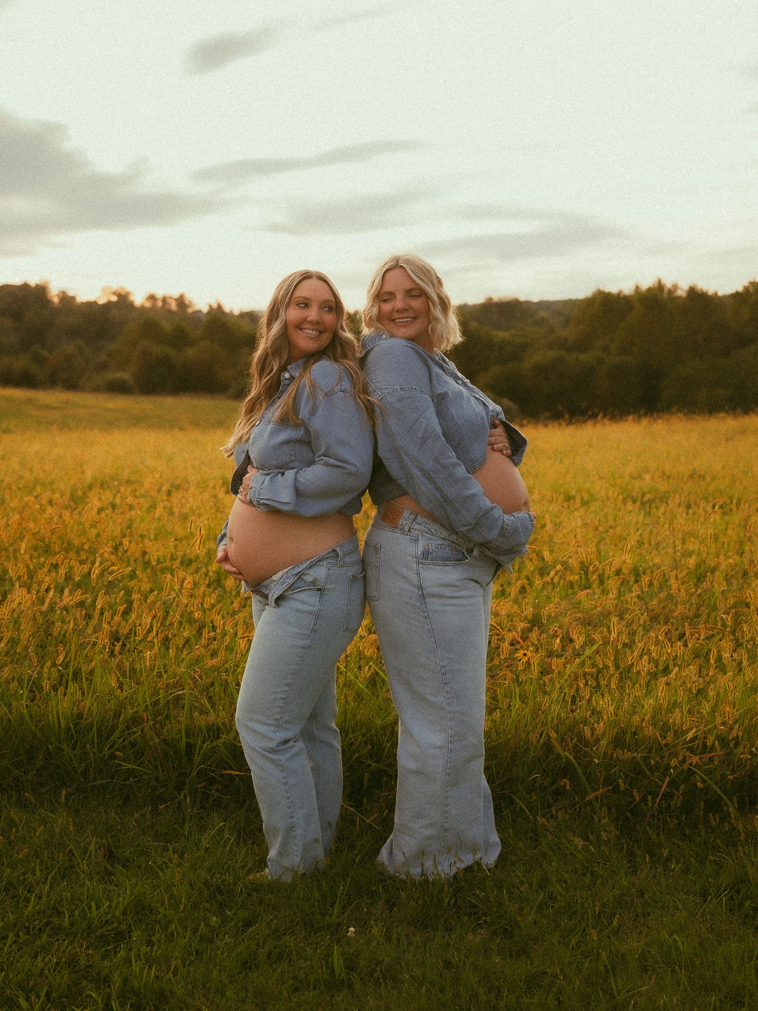Two pregnant women in denim outfits stand back-to-back in a golden field at sunset, smiling and showing their baby bumps.