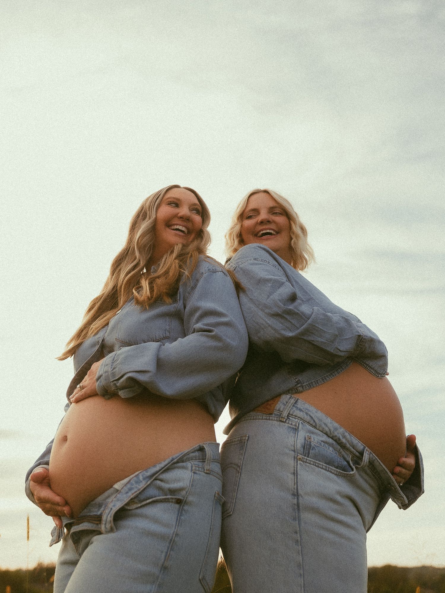 Two pregnant women in denim outfits pose back-to-back outdoors at sunset, smiling and cradling their baby bumps.