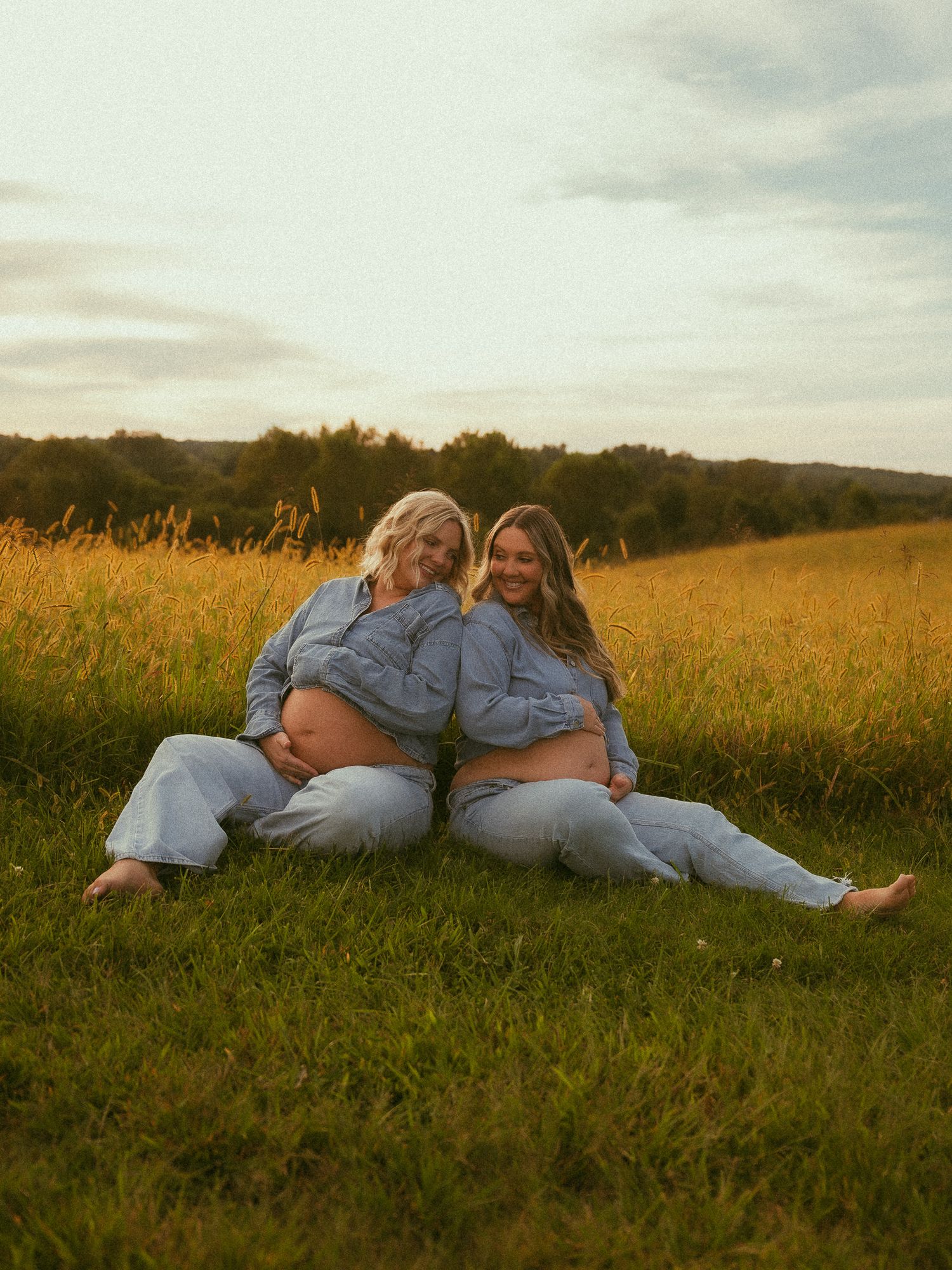 Two pregnant women in matching blue denim outfits sit back-to-back in a golden field at sunset.