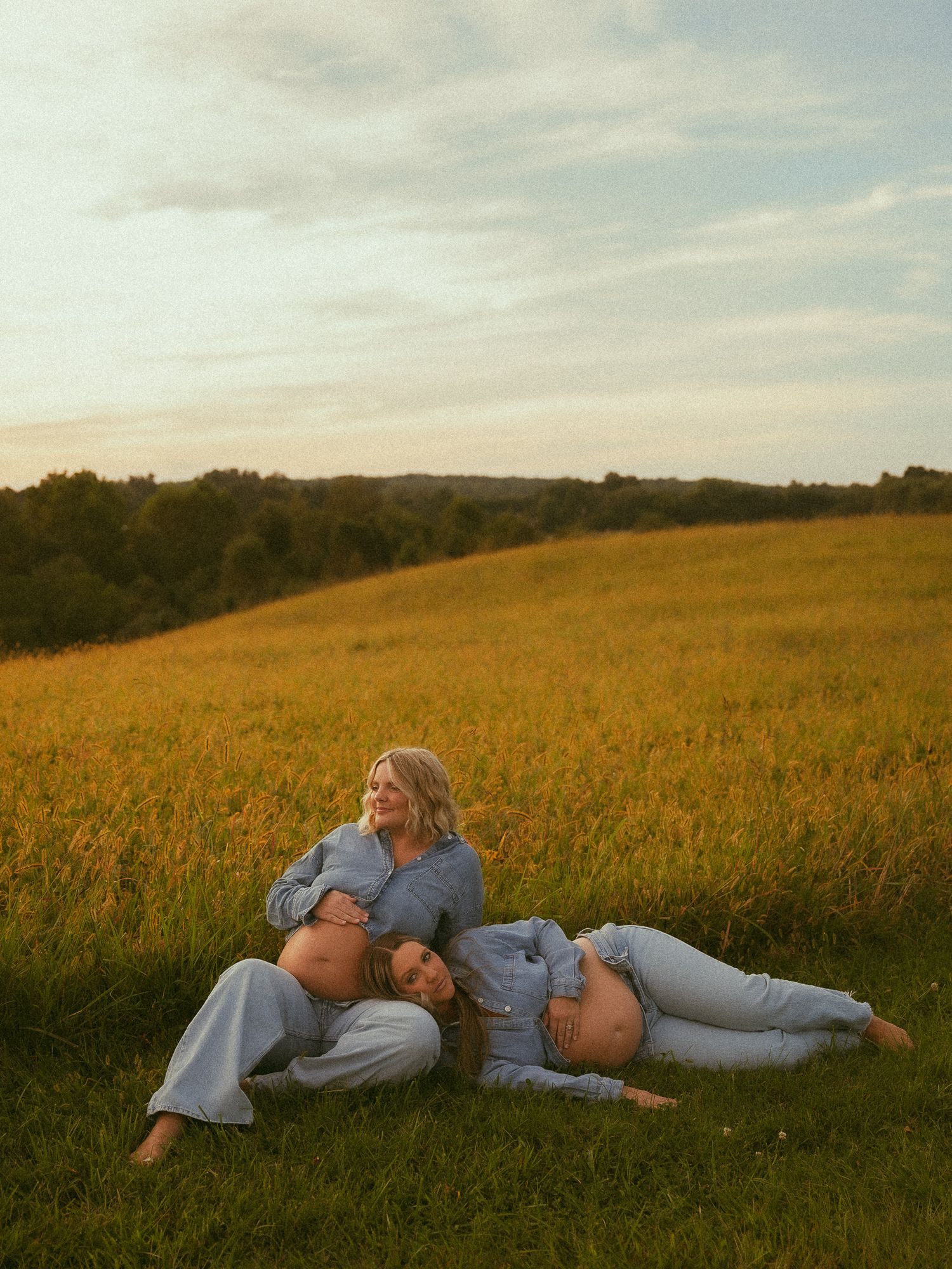 Two pregnant women in blue outfits pose together in a golden field at sunset during a maternity photoshoot.