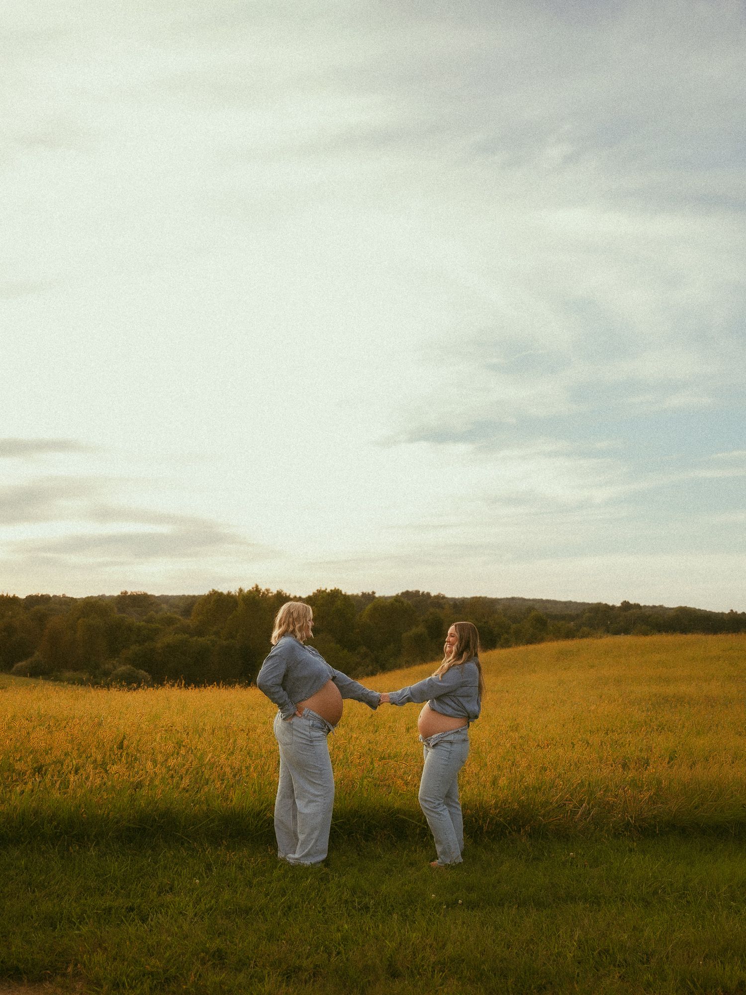 Two pregnant women holding hands in a golden field at sunset, wearing matching gray outfits during maternity photoshoot.
