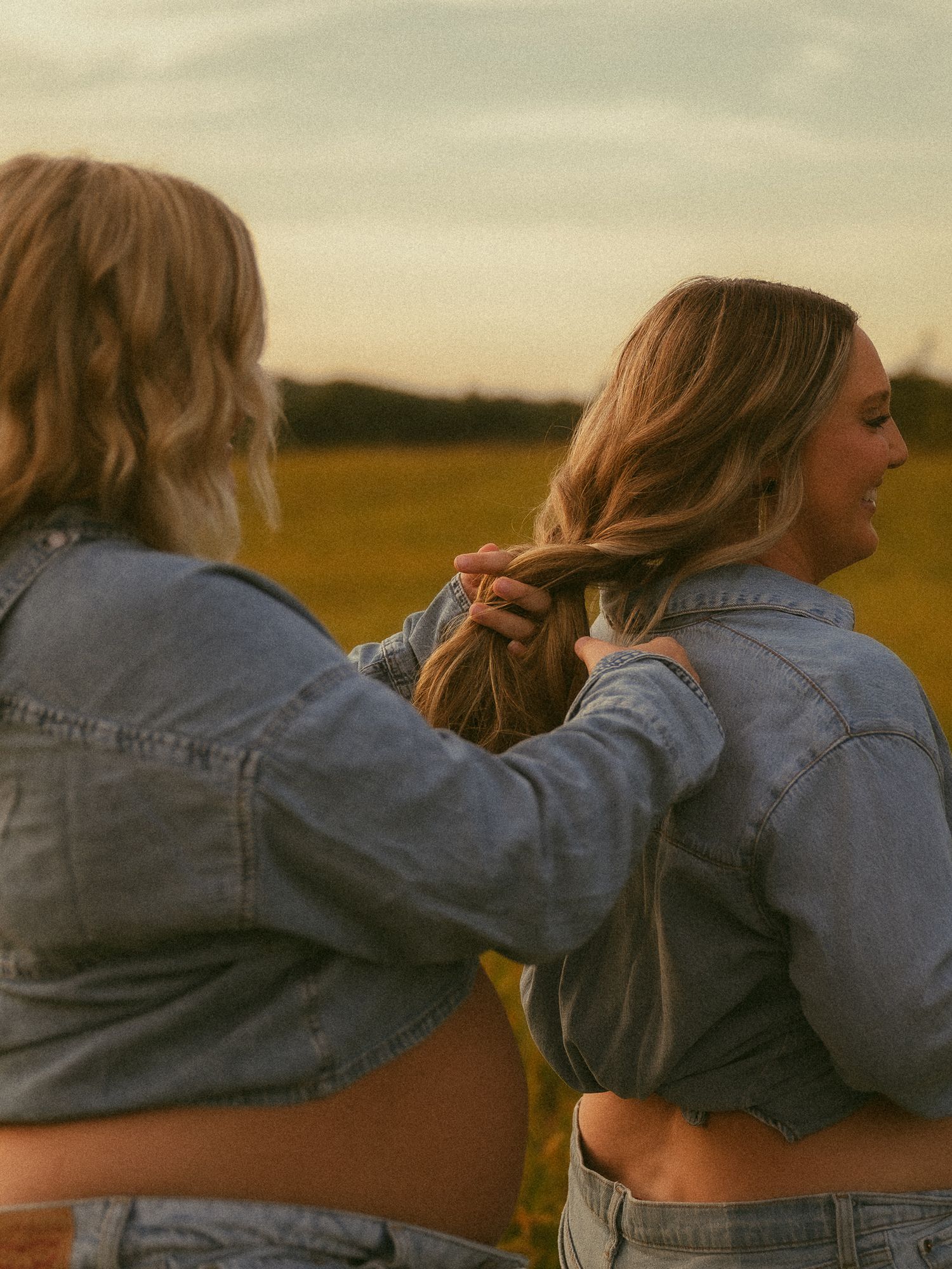 Two women in denim jackets braid hair outdoors at golden hour in a green field.