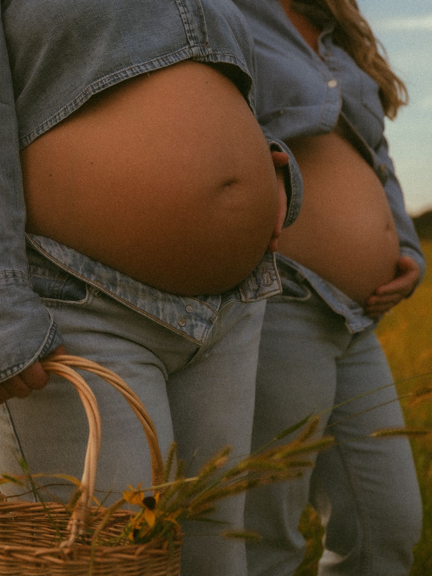 Two pregnant women in denim outfits stand outdoors holding a wicker basket with wildflowers in a field.