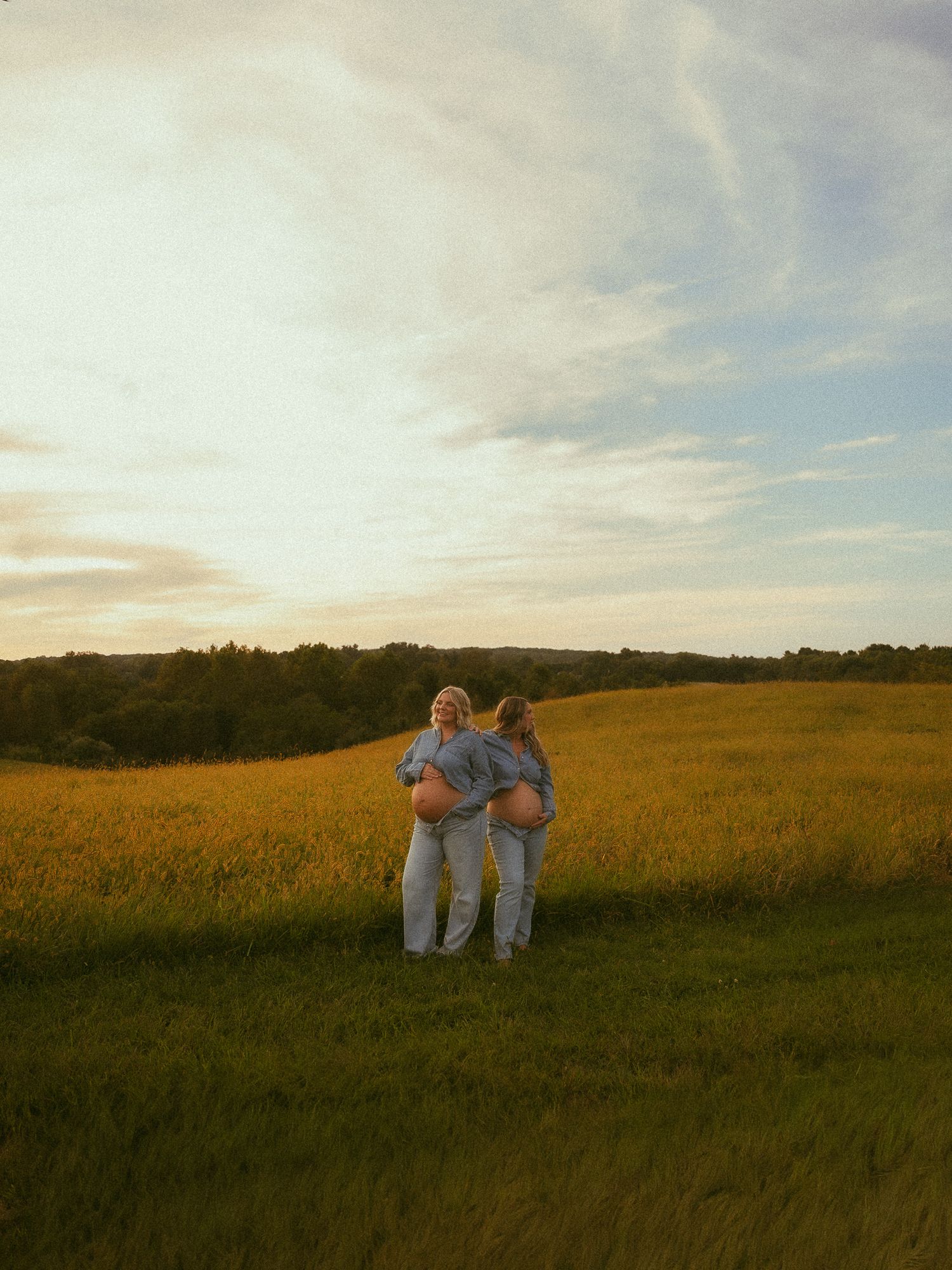 Two pregnant women stand back-to-back in a golden meadow at sunset, cradling their baby bumps in a serene outdoor setting.