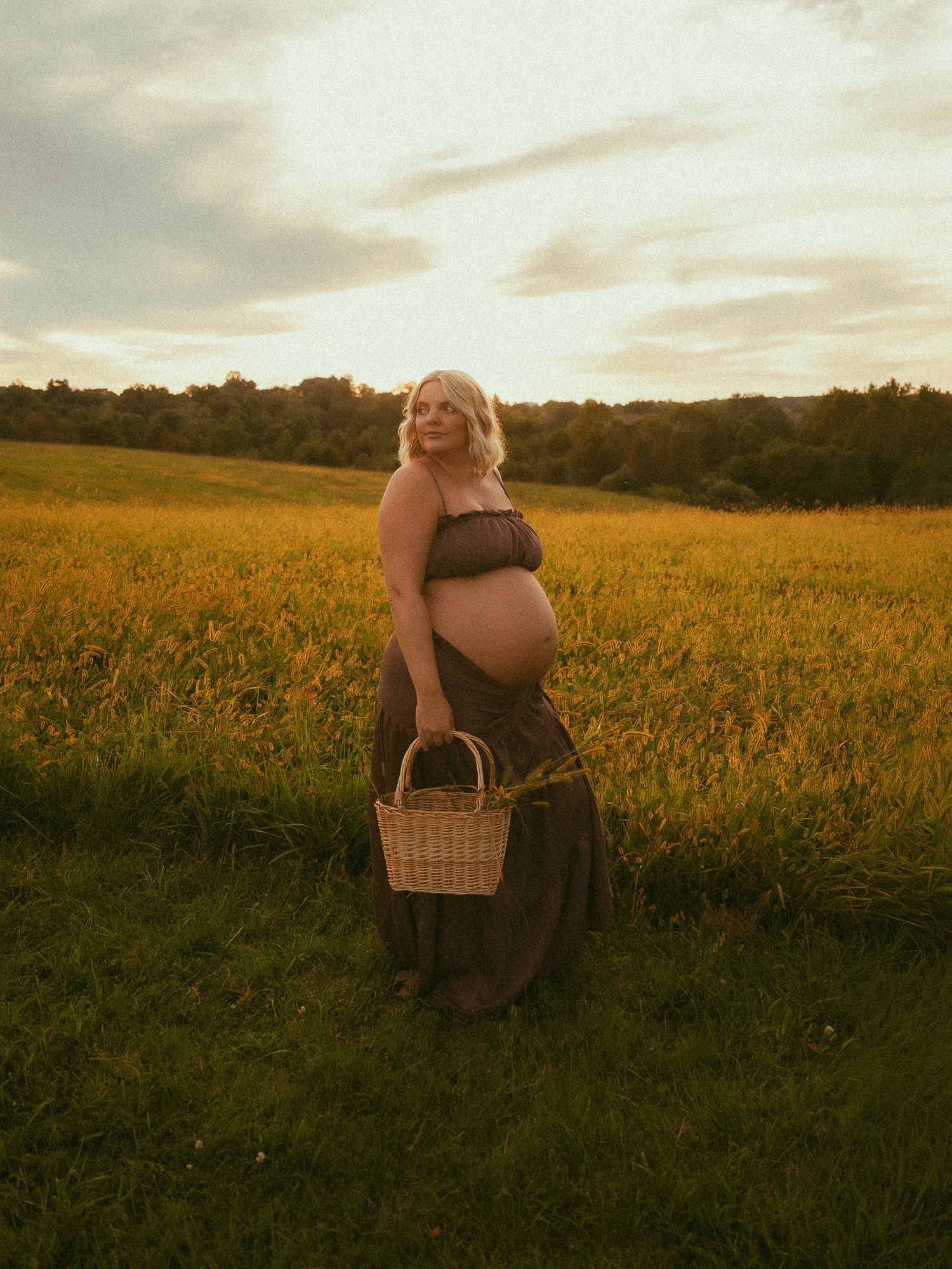 Pregnant woman in a dark maxi dress holding a wicker basket stands in a golden field at sunset.