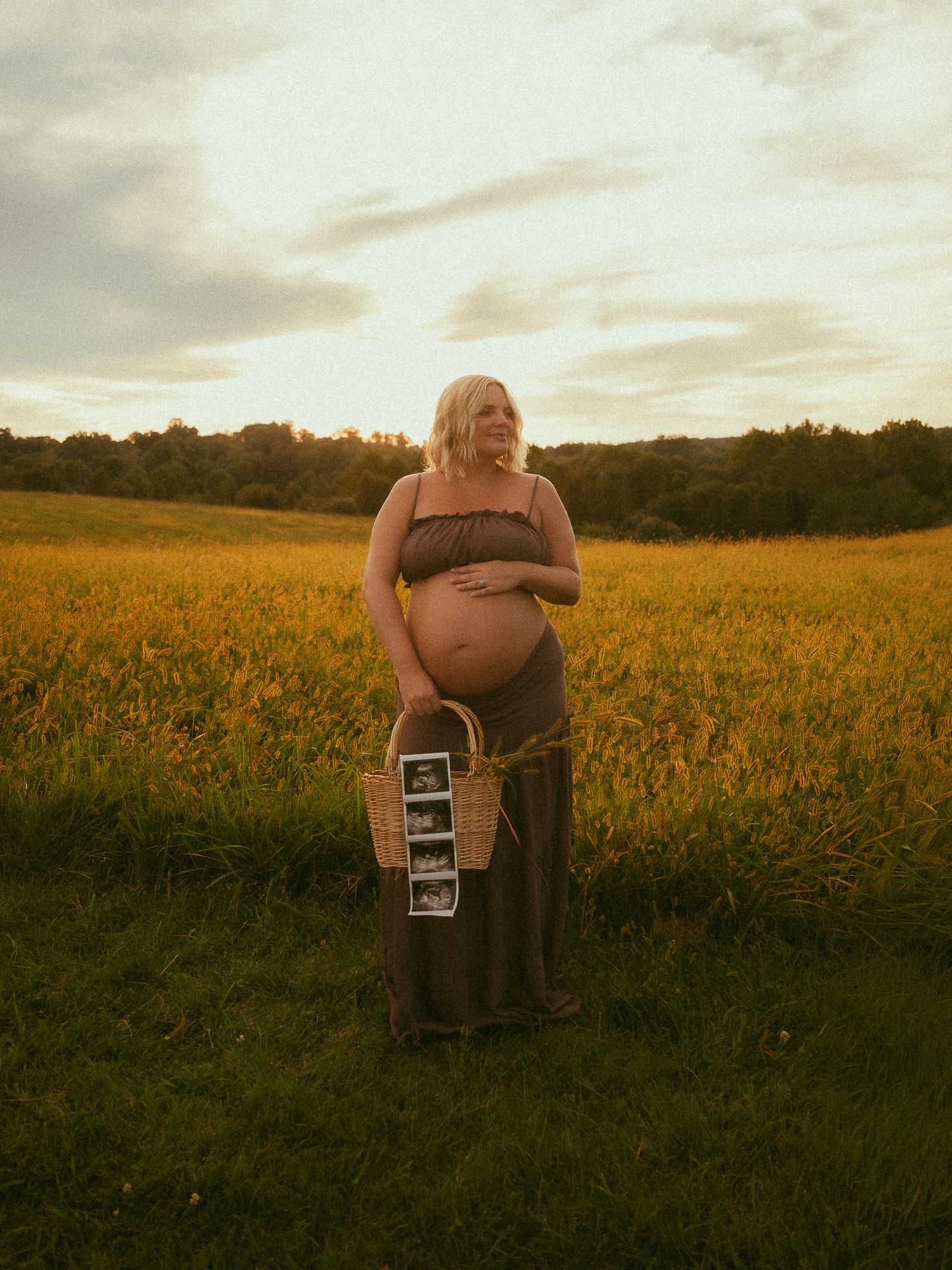 Pregnant woman in olive maxi skirt holds ultrasound photos in basket while standing in golden field at sunset.