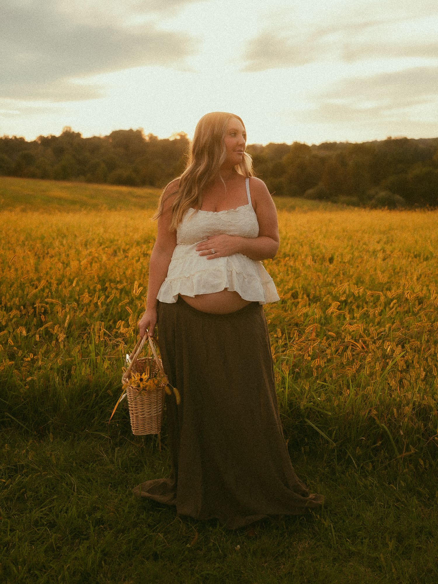 Pregnant woman in white crop top and olive maxi skirt holding a wicker basket in a golden sunlit field.