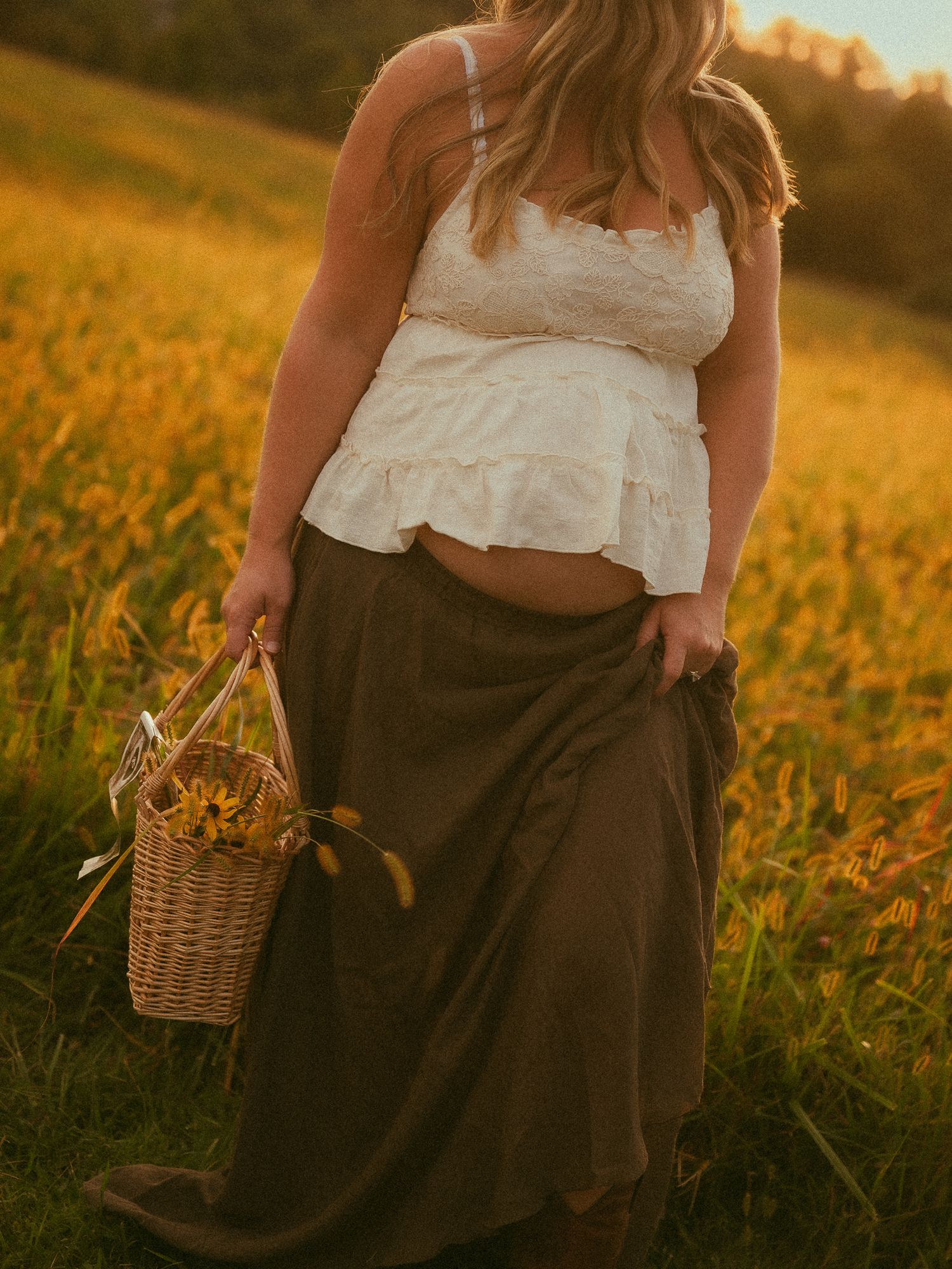 Woman in white lace crop top and olive maxi skirt holding a wicker basket with yellow flowers in a golden sunlit field.